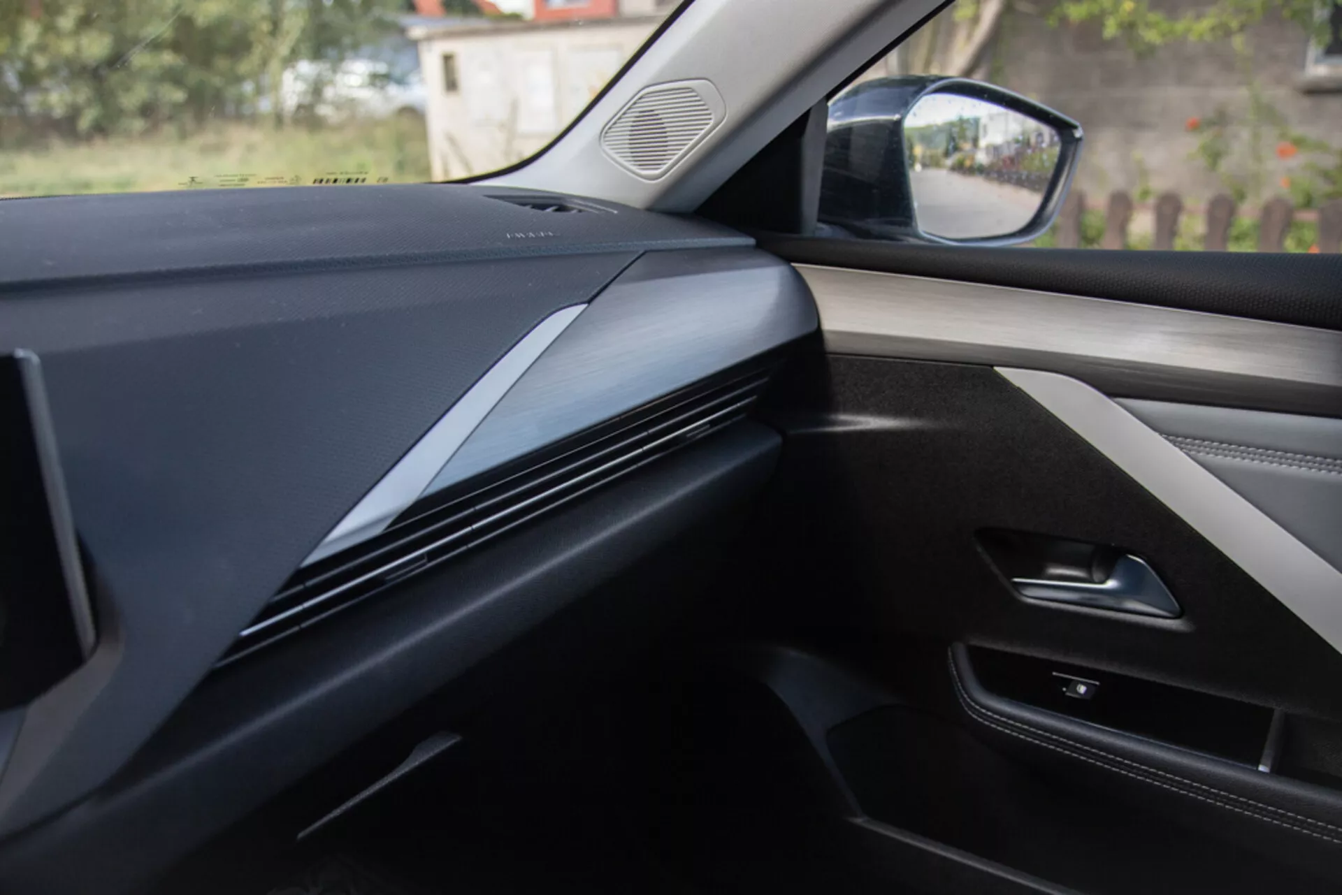Interiérový pohľad z perspektívy sedadla spolujazdca zobrazuje dizajn palubnej dosky, lišty a dverový panel v Opel Astra 2022, s tmavými, elegantnými kontúrovanými línia s dôrazom na textúru materiálu a potenciálne technologické integrácie