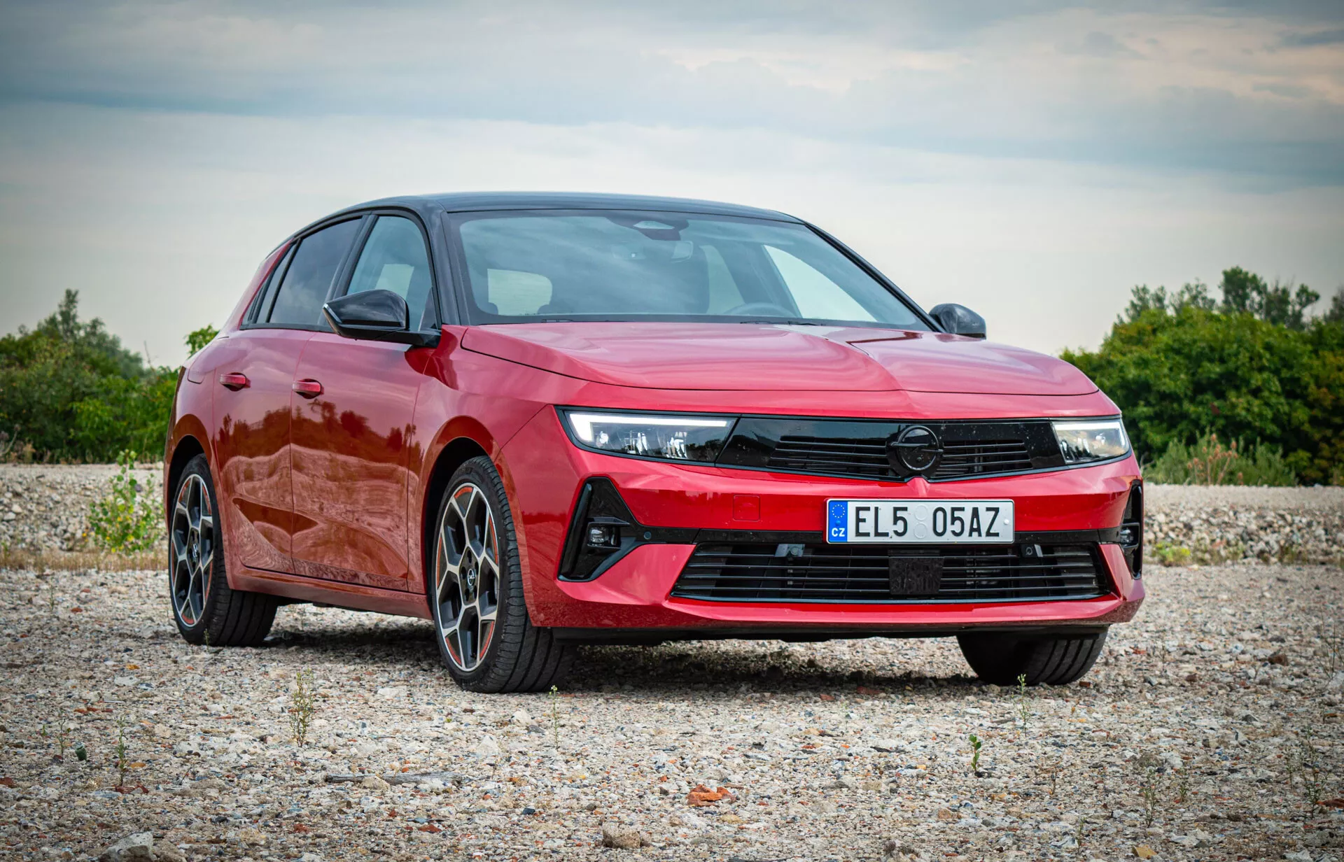 Opel Astra 2022 vo výraznej červenej farbe, zobrazuje moderný dizajn s uhlovými LED svetlami, výraznou prednou mriežkou, športovým predným nárazníkom a ľahkými zliatinovými kolesami s viacerými paprskami, predstavené v prírodnom vonkajšom prostredí z pohľadu z troch štvrtín spredu, čo zdôrazňuje aerodynamický profil a prémiové vlastnosti vozidla