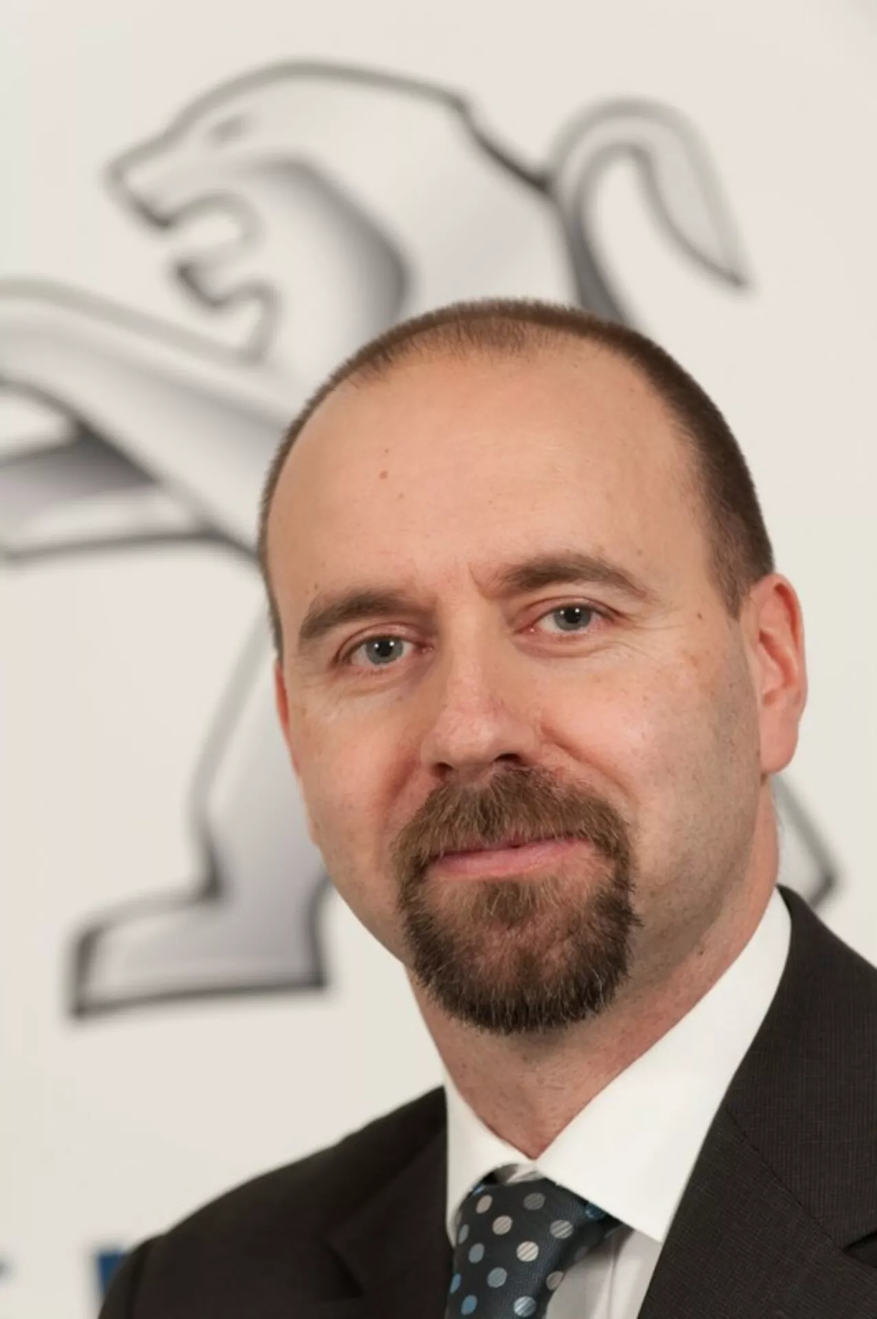 Profesionálny portrét muža v obleku a kravate s logom Peugeotu (lev) na pozadí, žiadne auto nie je viditeľné