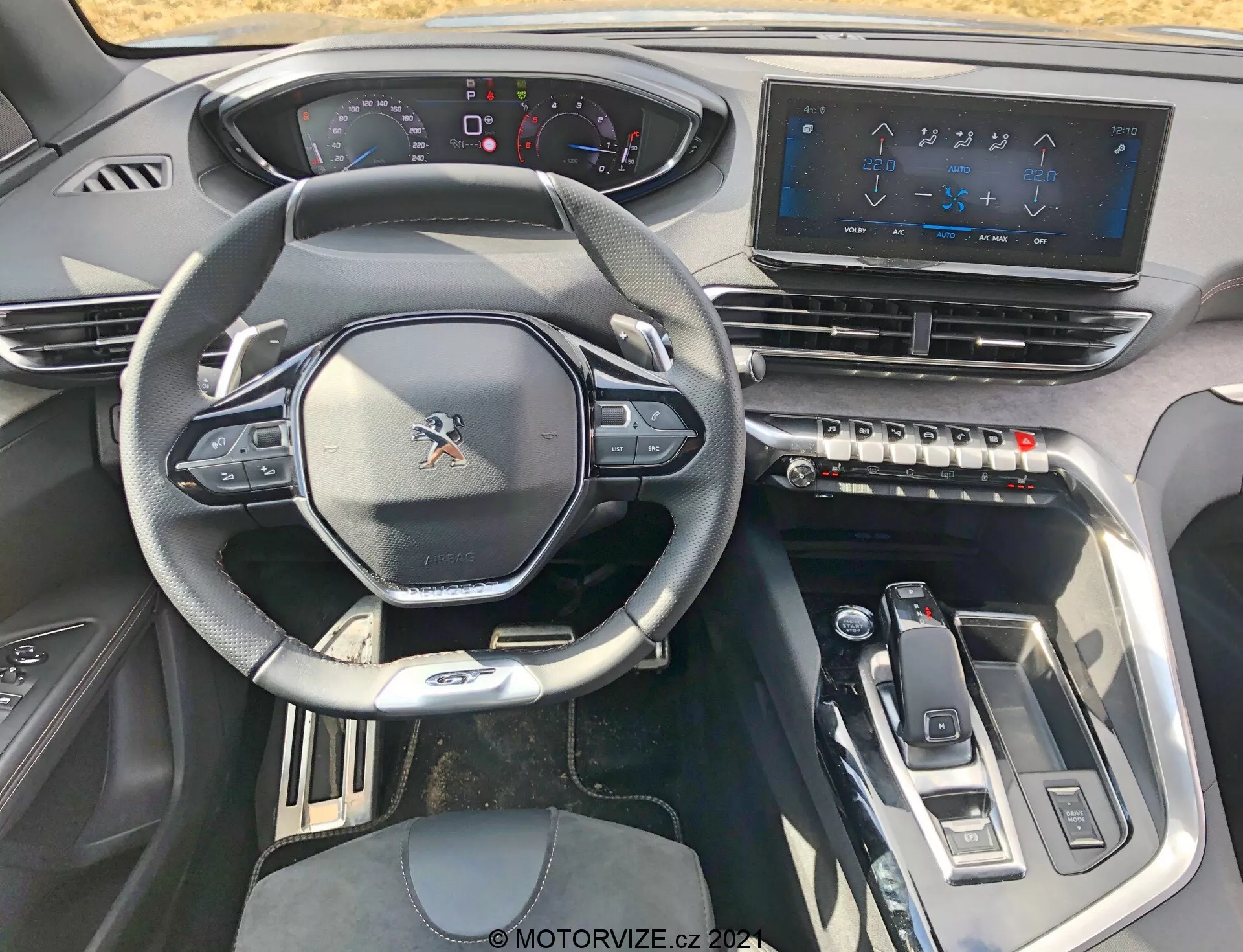 Pohľad do interiéru vozidla Peugeot 5008 z roku 2021 z pohľadu vodiča, ktorý ukazuje športový multifunkčný volant s ovládacími prvkami, čiastočne digitálny prístrojový panel, veľký dotykový infotainment systém, hmatové tlačidlá klimatizácie, elegantný volič prevodovky a moderné dizajnové prvky s kovovými akcentmi pre high-tech, luxusný zážitok.