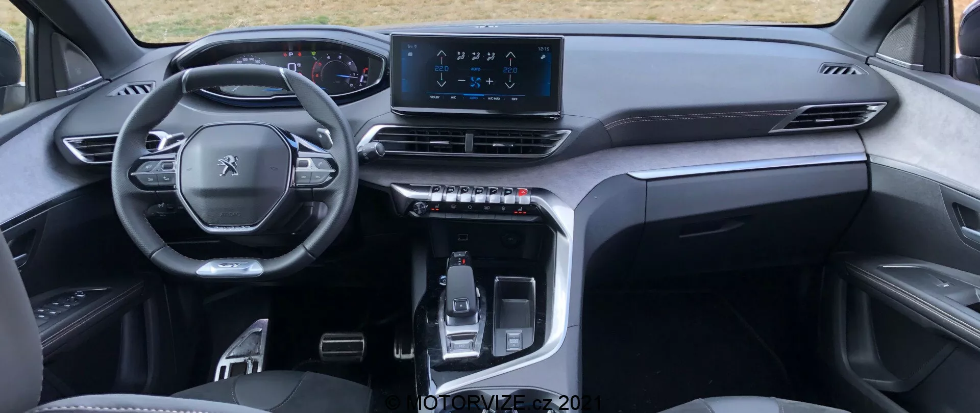 Pohľad na interiér Peugeot 5008 2021 z pohľadu zadného sedadla zdôrazňuje multifunkčný volant, digitálnu prístrojovú dosku, symetrickú palubnú dosku s ventilačnými otvormi, centrálnu dotykovú obrazovku infotainmentu a elegantnú stredovú konzolu s elektronicky ovládanou radiacej pákou a hmatovými tlačidlami. Štýlový minimalizmus s vysoko kvalitnými materiálmi naznačuje moderné, prémiové vozidlo.