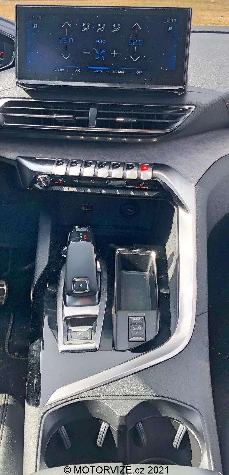 Pohľad do interiéru vozidla Peugeot 5008 z roku 2021, zameraný na stredovú konzolu z nadhľadu. Obrázok zvýrazňuje plávajúci infotainment dotykový displej zobrazujúci ovládanie klimatizácie nad fyzickými tlačidlami a ovládačmi pre rýchle nastavenie klímy. Nižšie je viditeľná elegantná elektronická radiaca páka a okolité ovládacie prvky pre výber jazdného režimu a multifunkčný ovládač. Dizajn naznačuje moderné, prémiové vozidlo s pokročilými technologickými a ergonomickými funkciami, vrátane držiakov na poháre a úložných priestorov.