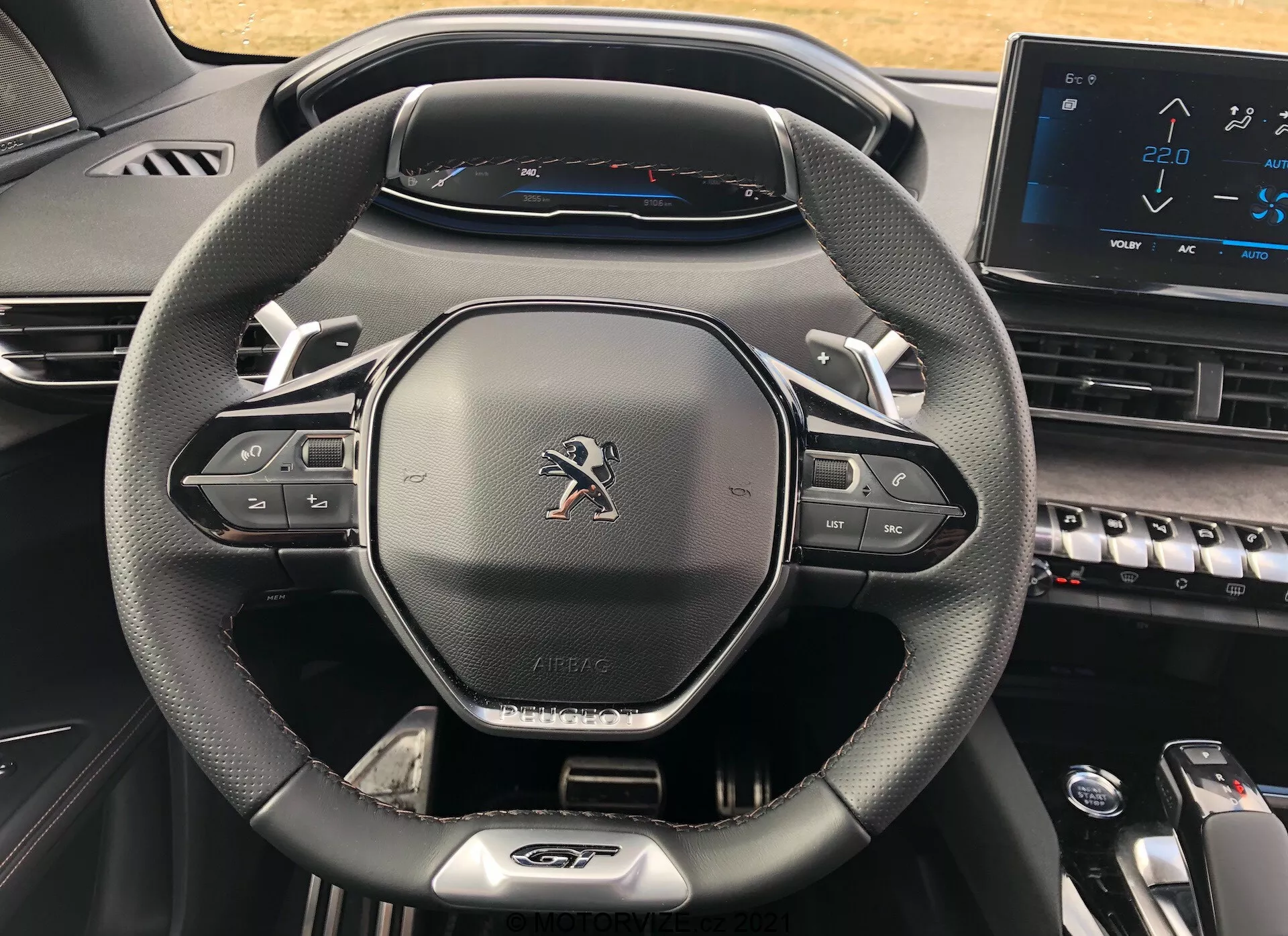 Detailný pohľad na kokpit vodiča v modeli Peugeot 5008 z roku 2021, zdôrazňujúci moderný trojramenný volant s koženým povrchom, tlačidlá na ovládanie multimédií a tempomatu, emblém Peugeot, označenie 'AIRBAG', plne digitálny prístrojový panel ukazujúci tachometer s maximálnou rýchlosťou 220 km/h a časť stredovej konzoly s klávesmi v štýle klavíra a elektronickým dotykovým displejom, čo predstavuje moderný a technologicky vyspelý dizajn.