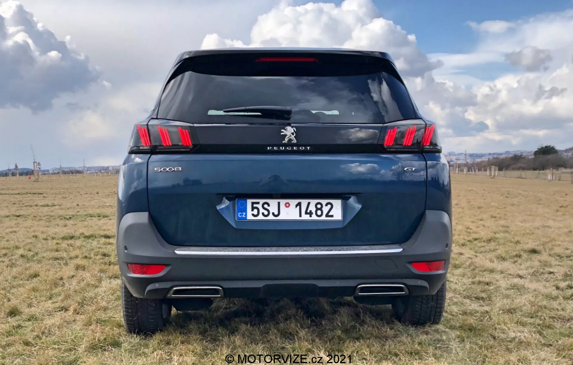 Pohľad zozadu na moderné SUV Peugeot 5008, zvýrazňujúci elegantné horizontálne koncové svetlá s charakteristickými LED segmentmi v tvare pazúrov, logo Peugeot a modelové označenie '5008'. Tmavomodrá alebo sivá farba, zaparkované na vonkajšom teréne s čiastočne zatiahnutou oblohou. Viditeľné prvky zahŕňajú zadné okno, zadný stierač, vysoko umiestnenú brzdovú sviečku, kľučku kufra a evidenčnú značku Českej republiky s 'CZ'. Reflexný pás nad nárazníkom so senzormi parkovania a výrezmi výfukov sú zreteľné. Fotka je centricky kompozíciovaná, vozidlo vyplňuje väčšinu rámu.