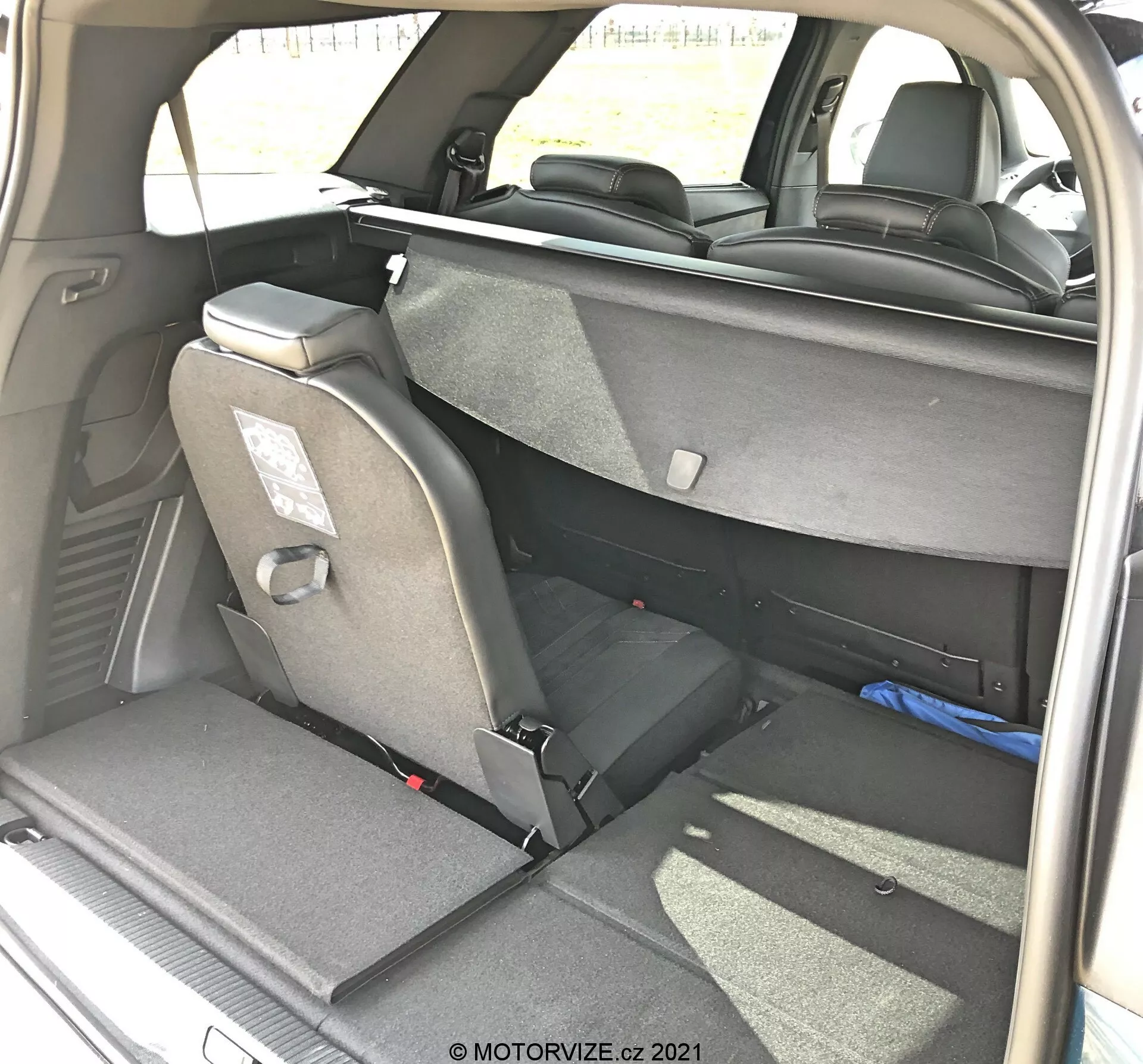 Pohľad na interiér nákladového priestoru Peugeot 5008 z roku 2021 z pravého zadného trojštvrťového uhla, ukazujúci flexibilné usporiadanie sedadiel s čiastočne sklopenými zadnými sedadlami, látkovým čalúnením, opierkami hlavy a bezpečnostnými pásmi. Pravé zadné sedadlo je úplne sklopené, ľavé sedadlo zostáva vo vzpriamenej polohe, čo naznačuje funkciu sklopenia sedadiel 60/40. Viditeľné tmavé vnútorné obloženie a koberec spolu s prirodzeným denným osvetlením, foto zhotovené MOTORVIZE.cz.
