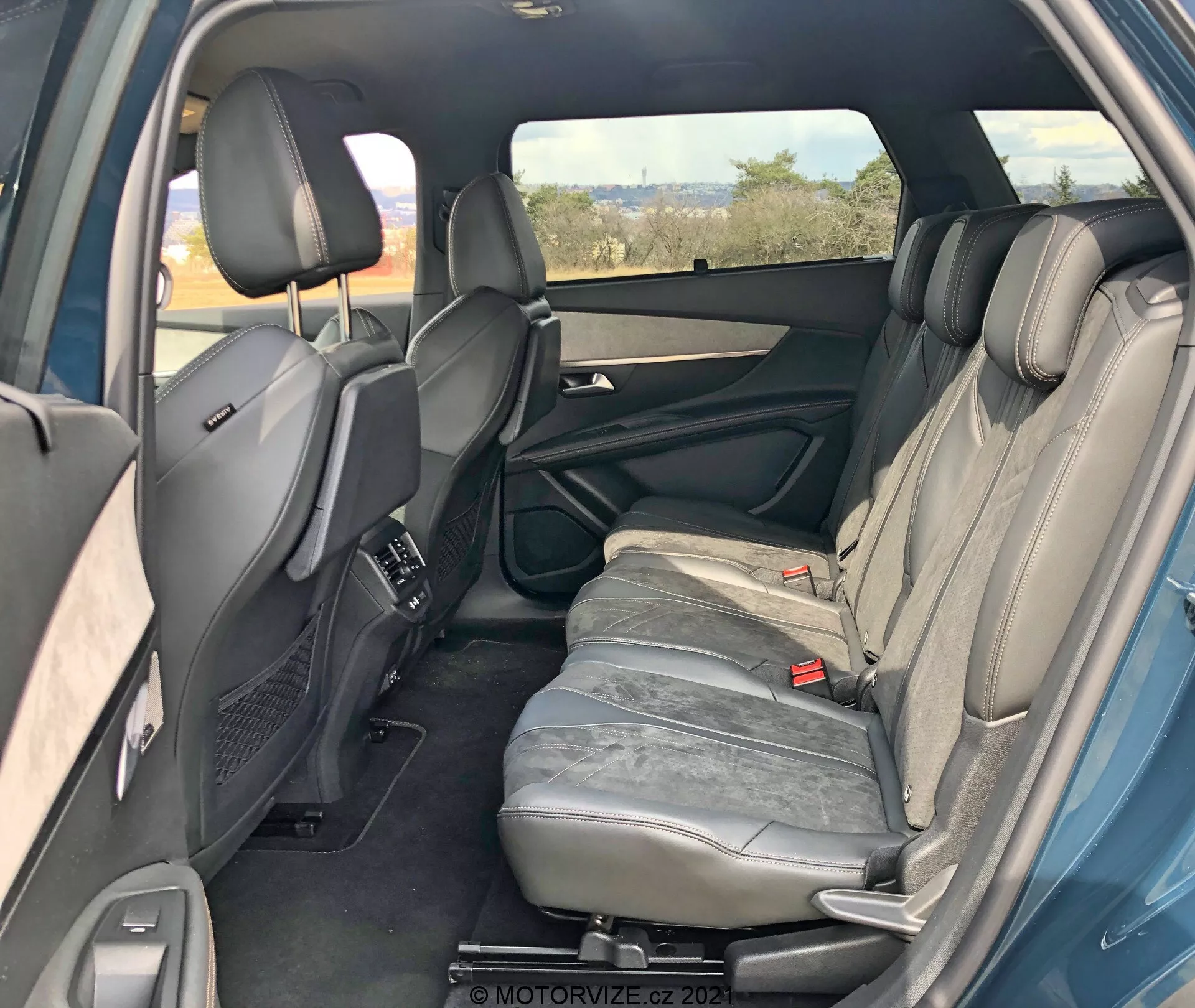 Interiér Peugeot 5008 2021 z perspektívy pravého zadného pohľadu, ktorý ukazuje zadný priestor pre cestujúcich. Pravé zadné dvere sú otvorené, čo odhaľuje prvky dverového panela, ako je kľučka, úložné priestory a reproduktor. Viditeľné sú tri zadné sedadlá čalúnené šedou kožou s opierkami hlavy. Stredná lakťová opierka sedadla je vo vzpriamenej polohe. Viditeľné sú tiež zadné bezpečnostné pásy a predné sedadlá s nastaviteľnými opierkami hlavy a stredovou konzolou. Zadné klimatizačné vetracie otvory na stredovej konzole, tónované pravé okno, náznaky ľavých zadných dverí naznačujúce symetriu. Celkovo dobre udržiavaný interiér bez viditeľných technologických funkcií.