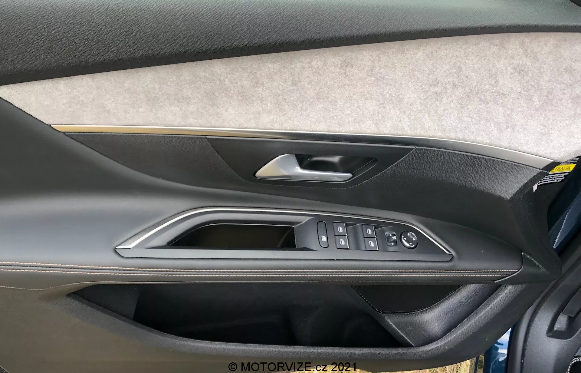 Detailný pohľad na vnútorný panel dverí Peugeot 5008 2021 s časťou čalúnenou filcom alebo textíliou, dvojfarebným dizajnom z tvarovaného plastu s tmavšou hornou časťou a svetlejšou spodnou časťou, vysoko kvalitnou kovovou alebo chrómovou kľučkou dverí, lešteným ozdobným prvkom a ergonomickými ovládacími prvkami pre elektrické okná, zámky dverí a bočné zrkadlá. Na spodnej časti panelu dverí je tiež viditeľné úložné vrecko.