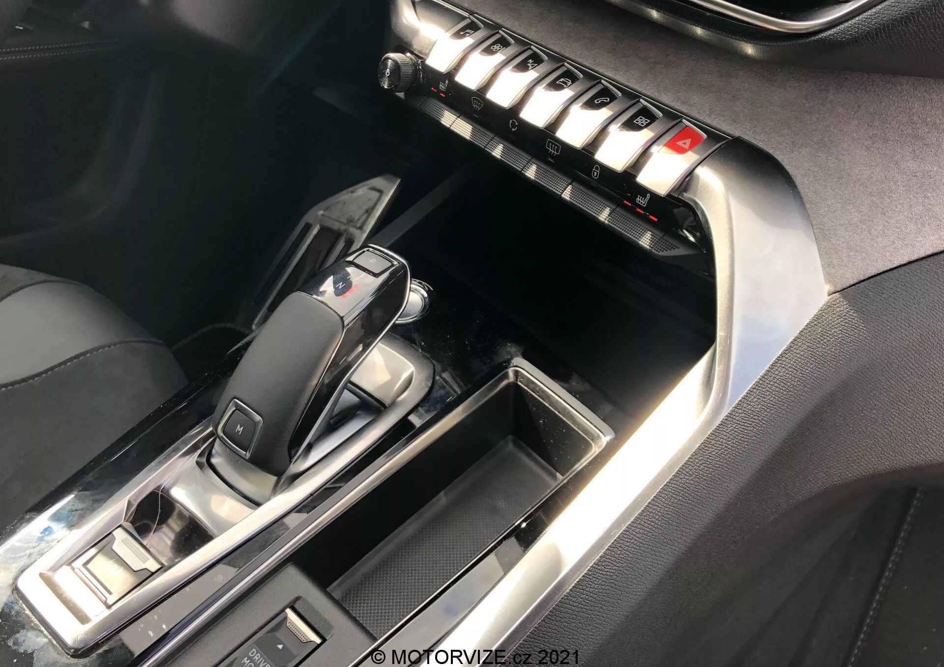 Pohľad zblízka na stredovú konzolu a predný interiér Peugeot 5008 2021, so zameraním na oblasť automatickej riadiacej páky a stredový panel. Páka je v polohe 'P', s elegantným dizajnom z lesklého čierneho plastu a kovovými prvkom. Okolité tlačidlá naznačujú ovládanie jazdných režimov a parkovacej brzdy. Stredový panel obsahuje rad kovových tlačidiel pravdepodobne pre ovládanie klimatizácie, odmrazovania a nastavenia sedadiel. Kontrastné prešívanie na koži alebo koženke a chromové akcenty zvyšujú luxusný pocit. Osvetlenie zvýrazňuje reflexné povrchy a detaily.