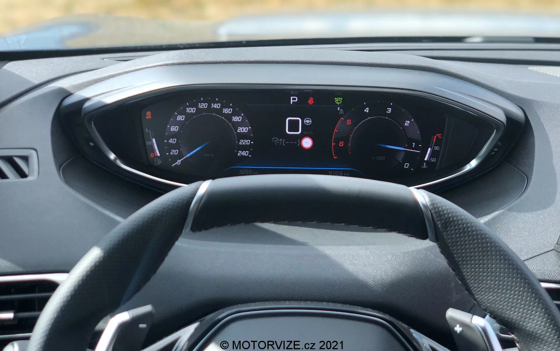 Pohľad na interiér moderného vozidla z pohľadu prvej osoby, zameraný na volant a digitálny prístrojový panel. Trojramenný volant je vycentrovaný, s rôznymi ukazovateľmi a indikátorom na osvetlenej prístrojovej doske za ním. Viditeľné sú tachometer a otáčkomer s hodnotami v km/h aj mph, vhodný pre medzinárodné trhy. Prístrojová doska zobrazuje množstvo varovných a informačných kontrolek, ako napríklad zapnutá parkovacia brzda, otvorené dvere, pripomienka bezpečnostného pásu a nízky tlak v pneumatikách, čo naznačuje pokročilé diagnostické a bezpečnostné funkcie. Farebná schéma prístrojovej dosky je modrá a červená na čiernom pozadí s bielym textom. Tento obrázok, nasnímaný za denného svetla, zdôrazňuje dizajn interiéru vozidla, digitálnu prístrojovú výbavu a modernú technológiu.