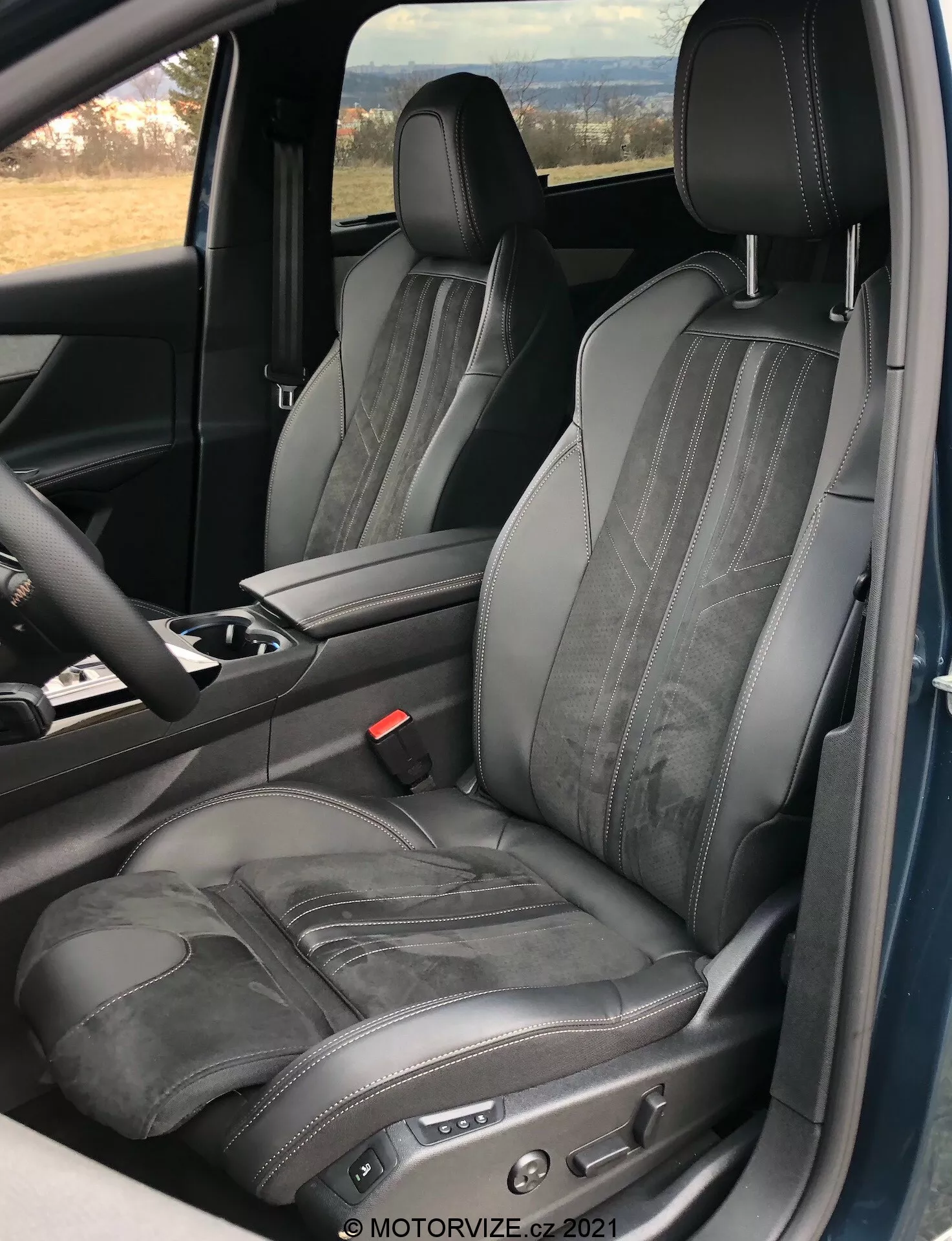 Pohľad na interiér vozidla Peugeot 5008 2021, zameraný na sedadlo vodiča, ukazujúci nastaviteľné čierne kožené sedadlo s kontrastným prešívaním a horizontálnym embosovaním, zhotovený z otvorených dverí, zdôrazňujúci luxusný a ergonomický dizajn s elektronickými ovládačmi nastavenia sedadla a viditeľnou časťou stredovej konzoly a volantu.