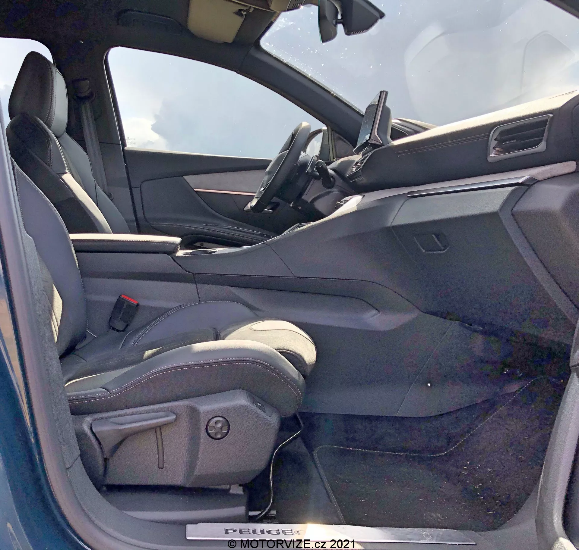 Pohľad na interiér Peugeot 5008 z roku 2021 z miesta vodiča s otvorenými dverami. Sedadlo vodiča je štýlu bucket, čalúnené pravdepodobne kožou alebo kožovitým materiálom, s bočnými límcami a ovládacími prvkami elektrického nastavenia. Otvorené dvere vodiča ukazujú vnútorný panel s ovládaním elektrických okien, pravdepodobne aj zrkadiel a mechanizmov zamykania dverí. Palubná doska obsahuje centrálne umiestnenú dotykovú obrazovku infotainmentu s minimalistickým dizajnom. Viditeľná časť vrchu volantu naznačuje kožený volant so zabudovanými ovládacími prvkami. Interiér má dvojfarebnú schému s tmavšími hornými povrchmi a svetlejšími spodnými povrchmi.