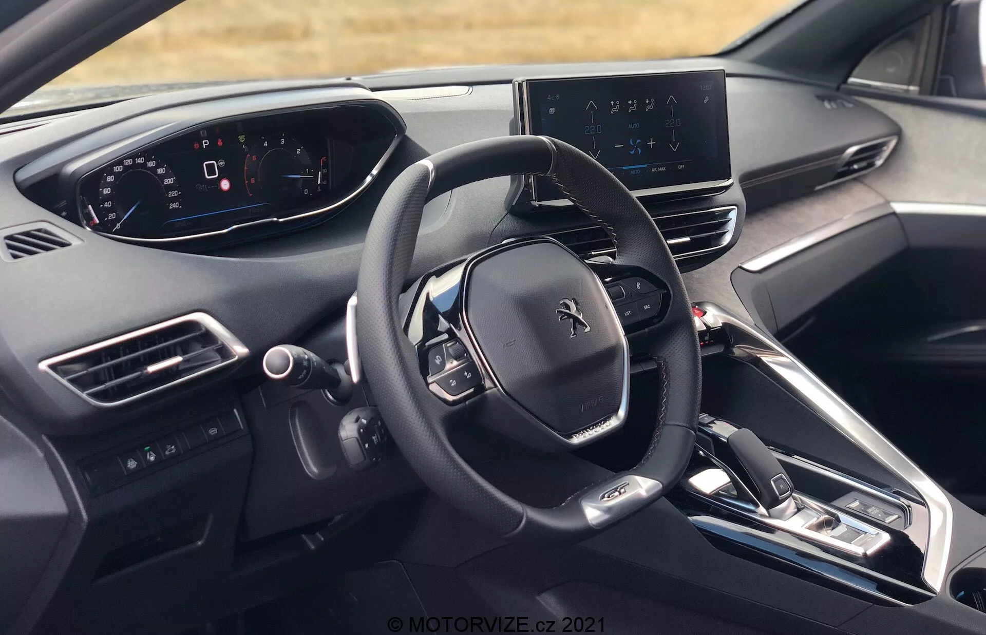 Pohľad na interiér vozidla Peugeot 5008 2021 z perspektívy vodiča, zobrazujúci volant s logom Peugeot, viacero ovládacích tlačidiel, plne digitálny prístrojový panel, centrálny infotainment displej s navigačnou mapou, elegantnú palubnú dosku s minimom fyzických tlačidiel, stredovú konzolu s tlačidlami na ovládanie klimatizácie a nastavenia vozidla, elektronickú radiacu páku, ventilačné otvory, tlačidlo štart/stop motora, materiály vyššej triedy a čiastočný pohľad na opierku hlavy vodiča.