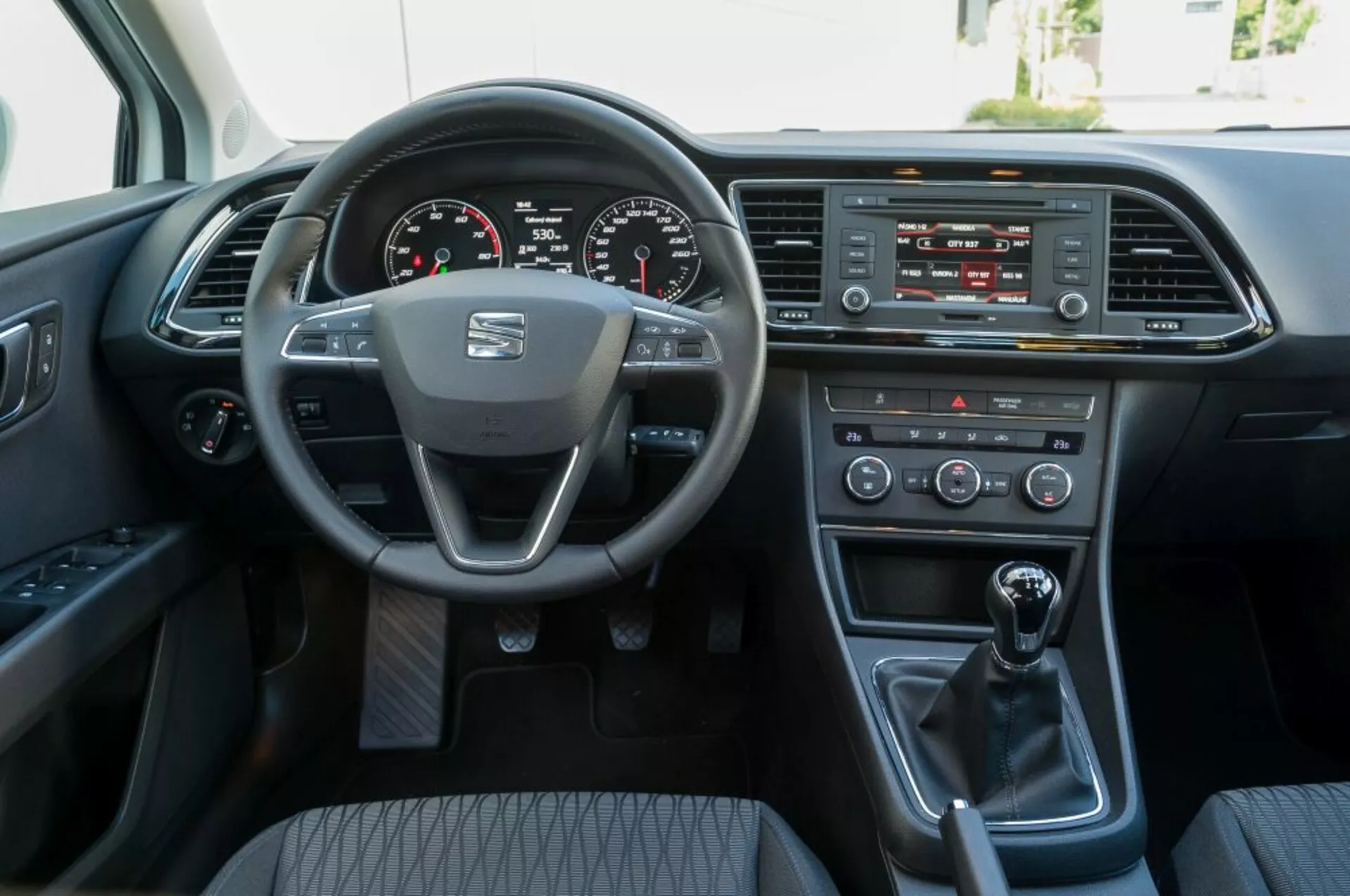 Interiérový pohľad na Seat Leon SC z roku 2016 z perspektívy predného spolujazdca, ukazujúci volant s logom výrobcu a namontovanými ovládacími prvkami, digitálny prístrojový panel, systém infotainment, ovládanie klímy, manuálnu radiacu páku a dvere vodiča s elektrickými možnosťami.