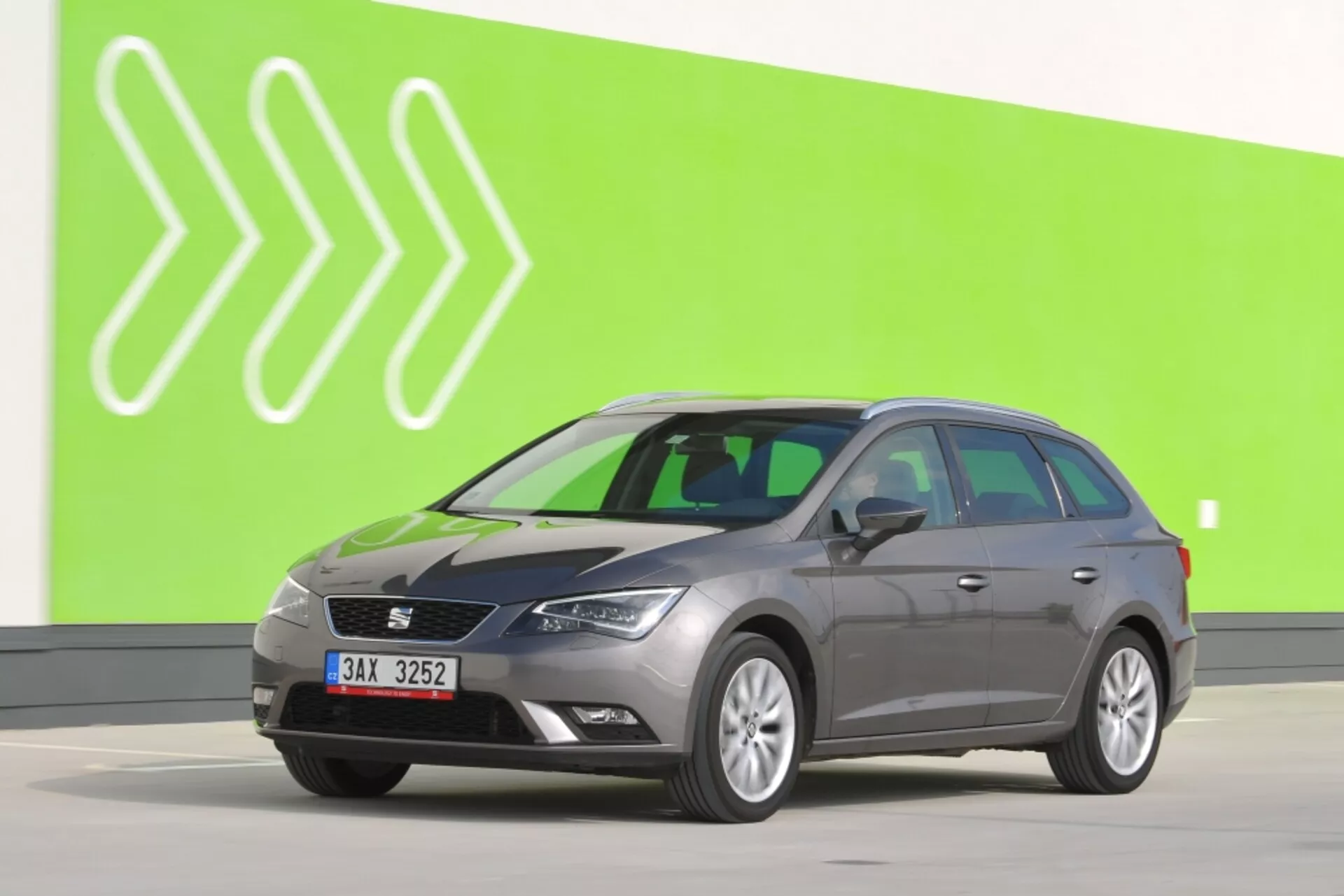 Šedé kombi Seat Leon SC 2012 v plnom bočnom pohľade na zelenom pozadí so štylizovanými bielymi šípkami, ukazujúce bočné zrkadlá s blinkry, charakteristické kontúry dverí a štandardné kolesá na svetlosivej podlahe štúdia
