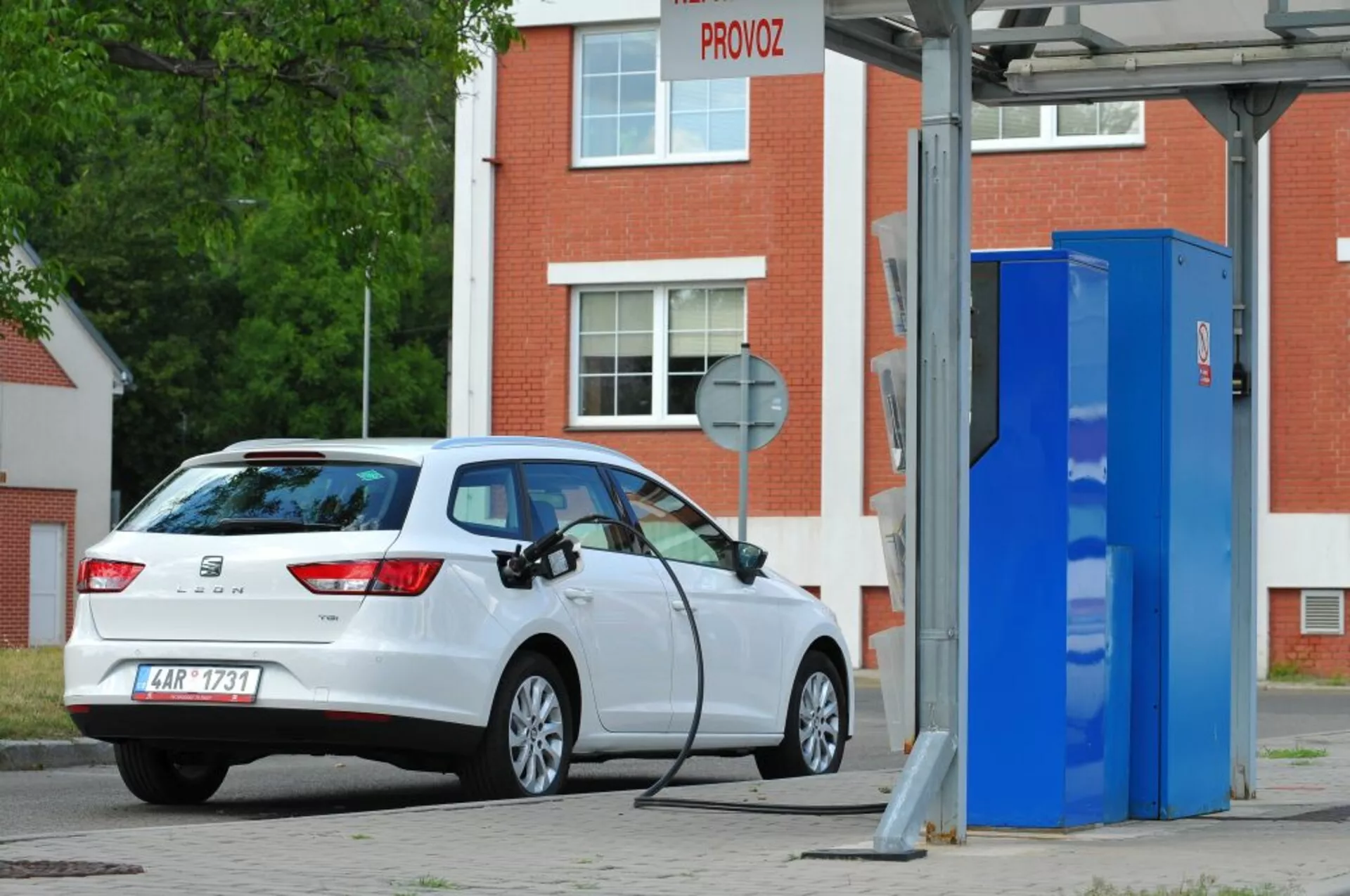 Biely hatchback Seat Leon SC z roku 2016, elektrický alebo plug-in hybrid, zaparkovaný pri nabíjacej stanici pre elektromobily, zobrazujúci pohľad z trojštvrťovej zadnej časti, ktorý zdôrazňuje zadné svetlá, zadné čelné sklo, profil zadnej časti a nabíjací port na pravej strane