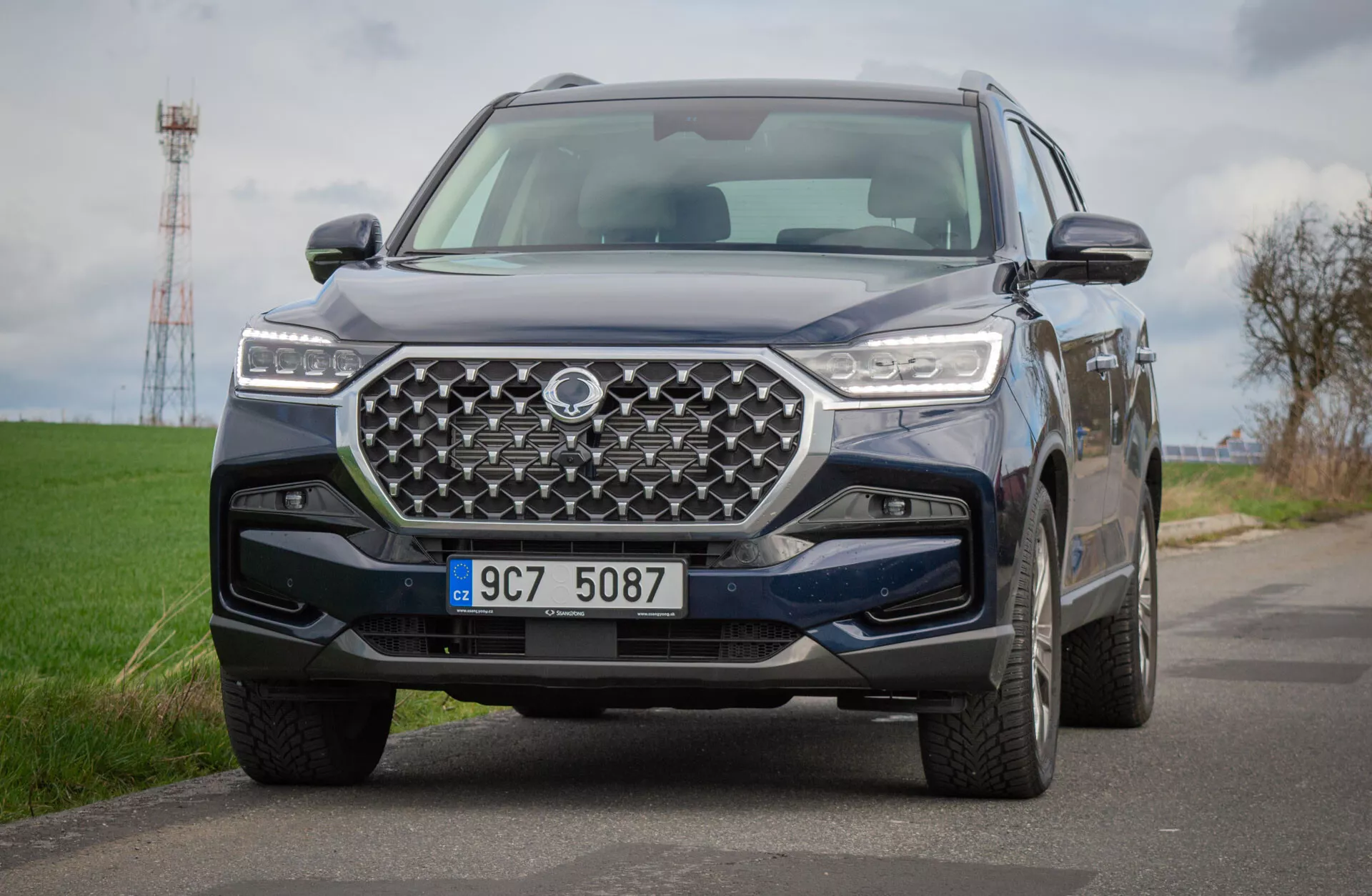 SUV SsangYong Rexton 2021 na vidieckej ceste, prezentujúce detailný pohľad na jeho predný dizajn s geometrickou mriežkou a LED svetlometmi, odzrkadľujúce pokročilú technológiu a schopnosti v teréne