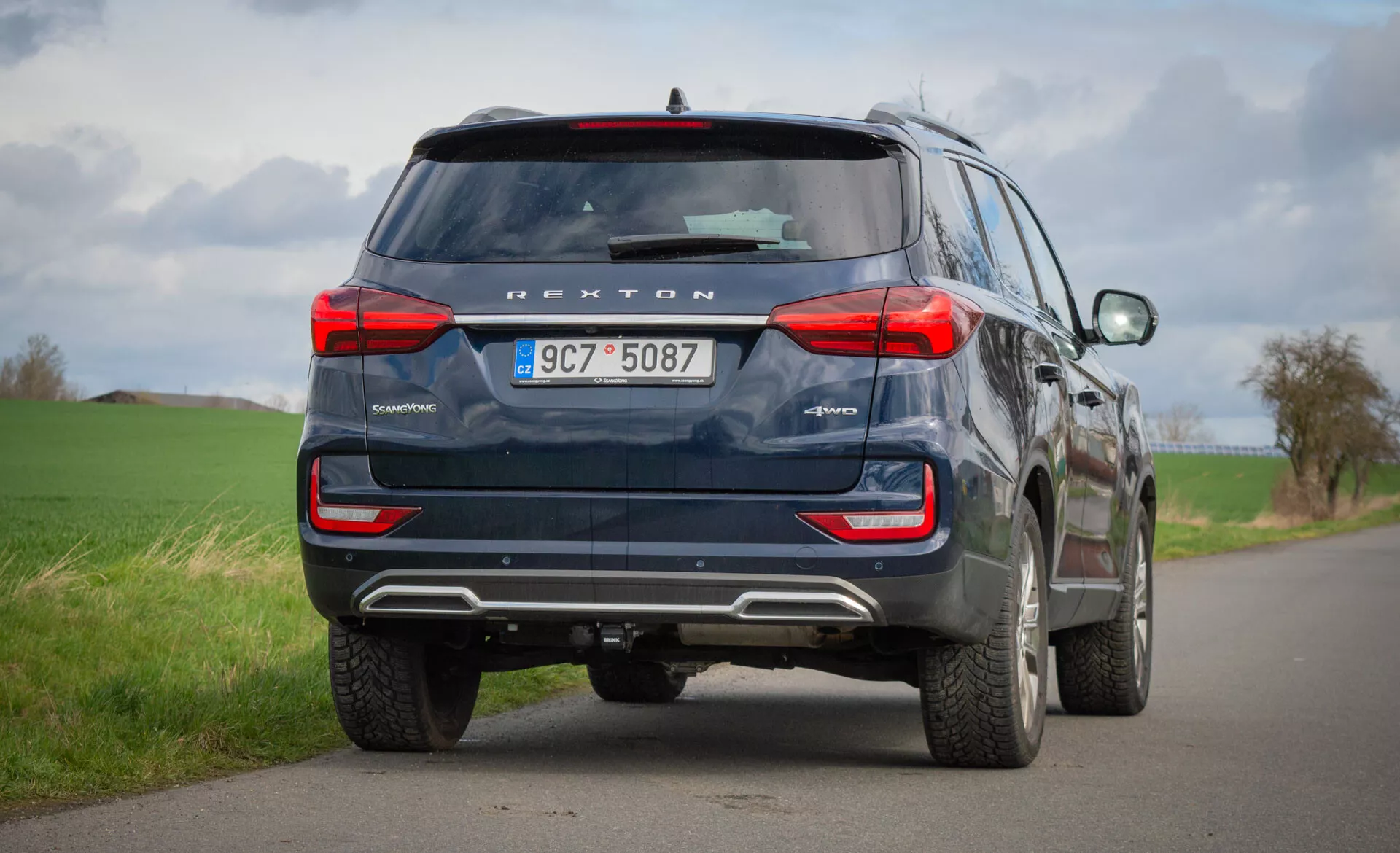 Zadný pohľad na modré SUV SsangYong Rexton 2021 zaparkované na vidieckej ceste s dvoma pruhmi, zdôrazňujúce jeho dizajnové prvky ako sú zadné svetlá, zadné okno, výfukové rúry a bezpečnostnú technológiu vrátane kamery na zadnom pohľade a parkovacích senzorov pod zamračenou oblohou.