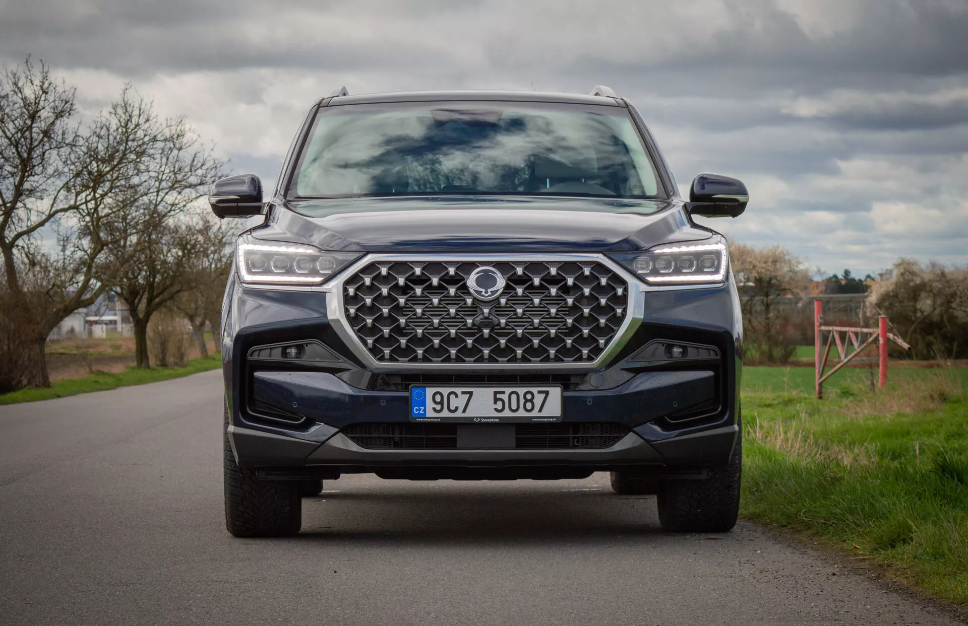 Moderné SUV SsangYong Rexton 2021 vo farbe, zachytené z pokľačanej pozície zobrazujúcej prednú časť vozidla, LED svetlomety, veľká dizajnová mriežka, predný nárazník so spodnou ochranou a pokročilé asistenčné systémy vodiča