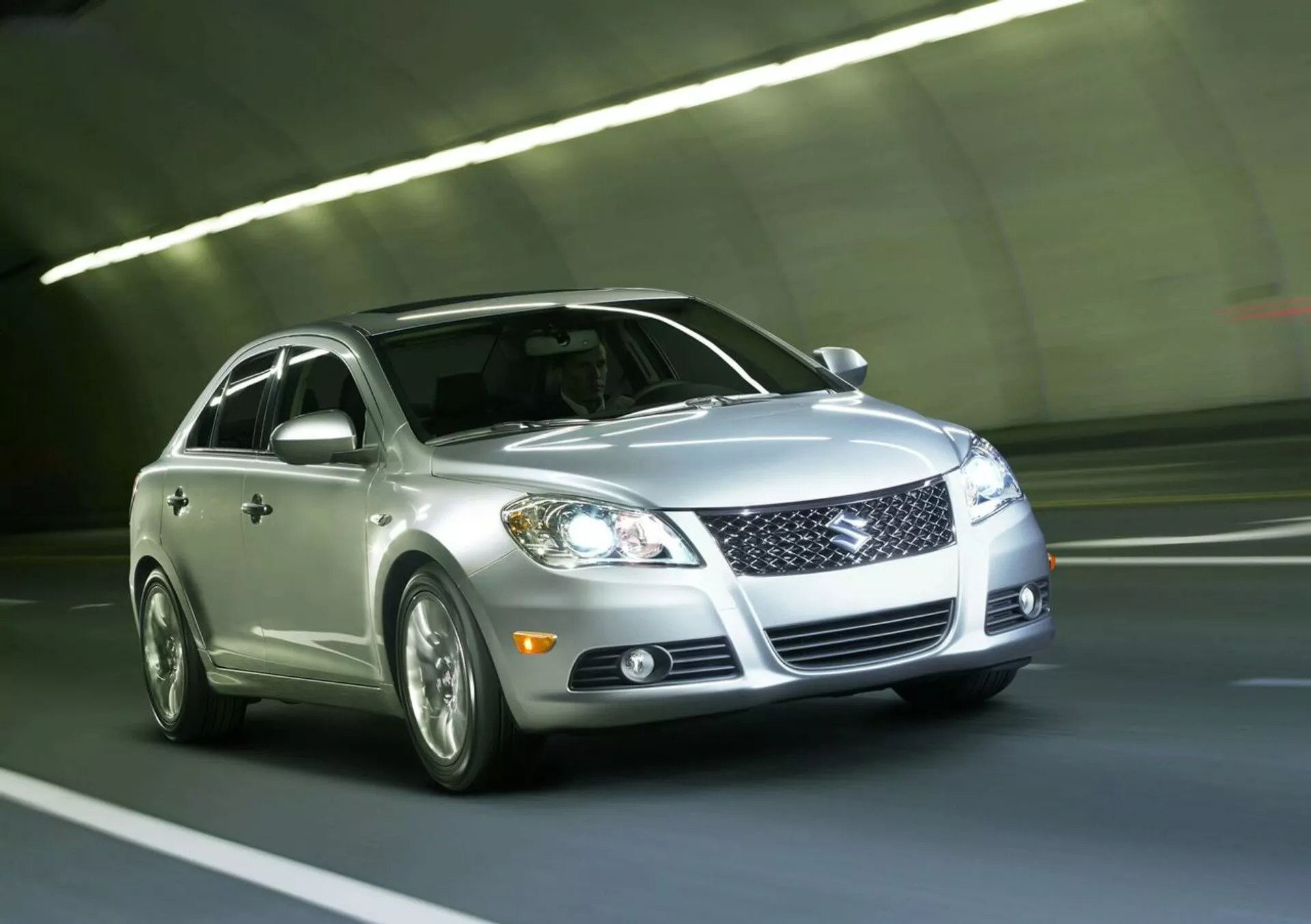 Strieborný sedan Suzuki Kizashi 2010 prechádzajúci tunelom, zobrazujúci prednú mriežku, LED svetlomety, hmlové svetlá, zliatinové kolesá a odrazivý lak, pohľad z nízkeho predného trojštvrtečného uhla zdôrazňujúci dynamický dizajn a technológiu vozidla.