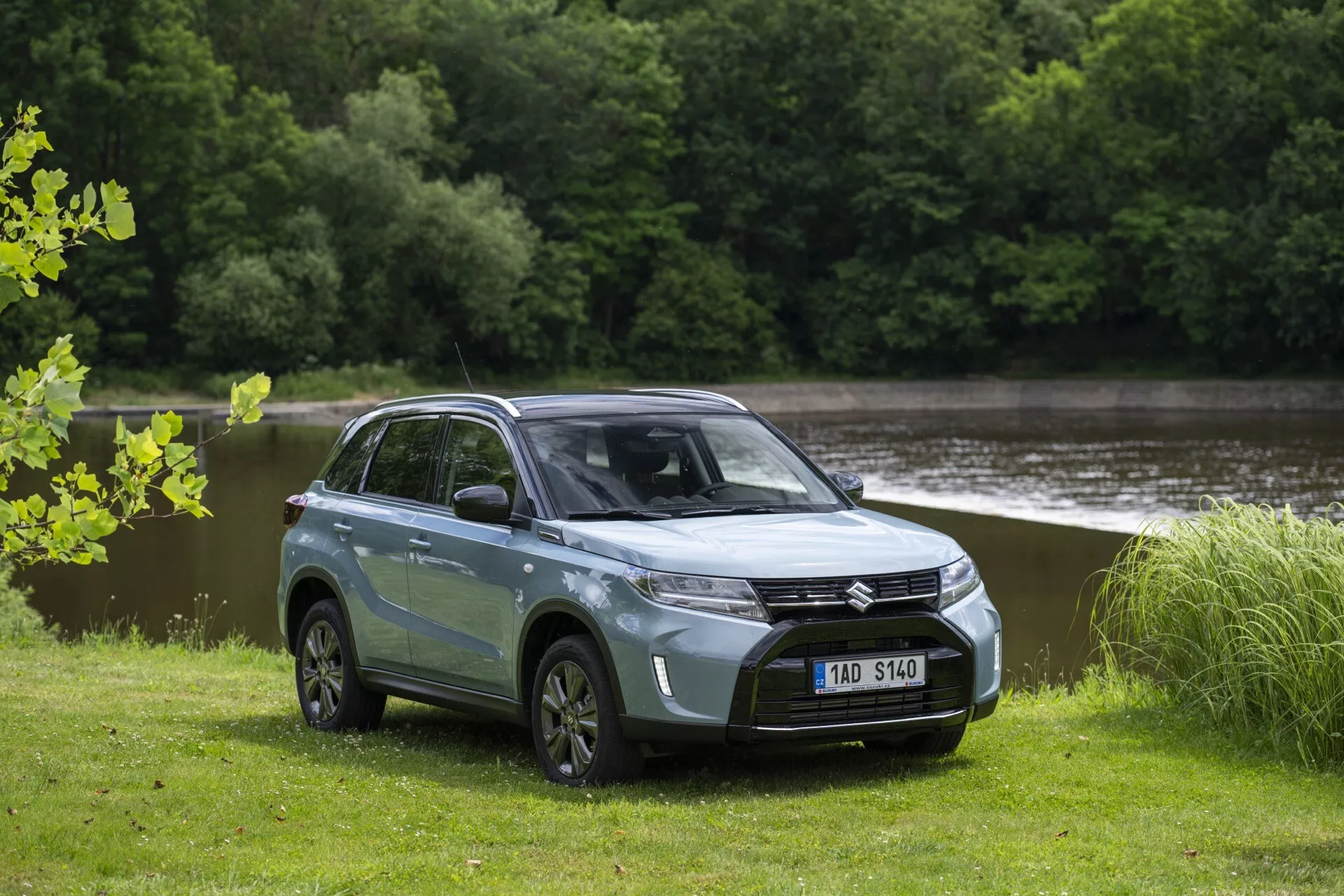 Fotografia moderného SUV Suzuki Vitara 2024 (2. generácia, facelift 2) v dvojfarebnej karosérii, svetlo modrej s čiernou strechou, zaparkovaného na trávnatom poli s vodou a stromami v pozadí. SUV je zobrazené z predného kvadrantového uhla, ukazujúc na stranu vodiča. Má výraznú prednú mriežku s centrálnym emblémom, elegantné uhlové LED svetlá, hladkú kapotu, viaclúčové kolesá z ľahkých zliatin a výraznú svetlú výšku. Tónované okná, bočné zrkadlá a farebne zladené kľučky dverí zvýrazňujú moderný dizajn, zatiaľ čo bočné zrkadlá integrujú LED ukazovatele smeru.