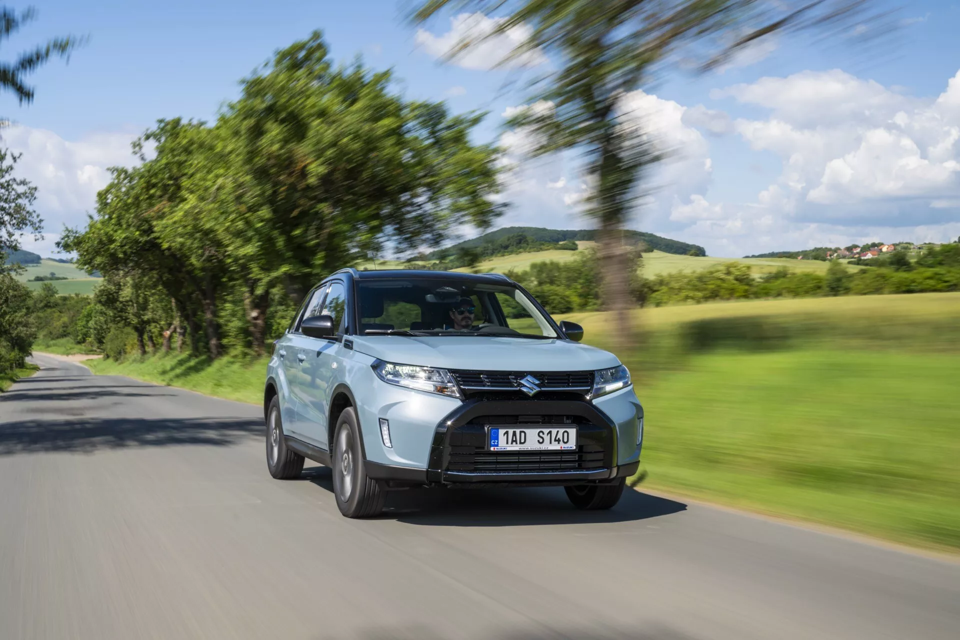 Suzuki Vitara 2024 Facelift 2. generácie je zachytený v pohybe s rozmazaným pozadím stromov a oblohy, zvýrazňujúc jeho rýchlosť. Tento pohľad z predného šikmého uhla zvýrazňuje moderné SUV so zvýšenou svetlou výškou, charakteristickou mriežkou, LED dennými svetlami, hmlovými svetlami a prívodmi vzduchu. Bočné zrkadlá, aerodynamické telo a pohybujúce sa zliatinové kolesá zdôrazňujú jeho dizajn. Viditeľné senzory naznačujú prítomnosť pokročilých asistenčných systémov vodiča, ako je adaptívny tempomat a vyhýbanie sa kolíziám. Kovový lak, čistý vzhľad a jasná, ale anonymná poznávacia značka prispievajú k jeho estetike. Vozidlo je pozicionované na vidieckej ceste, čo ukazuje jeho všestrannosť pre rôzne typy jázd.