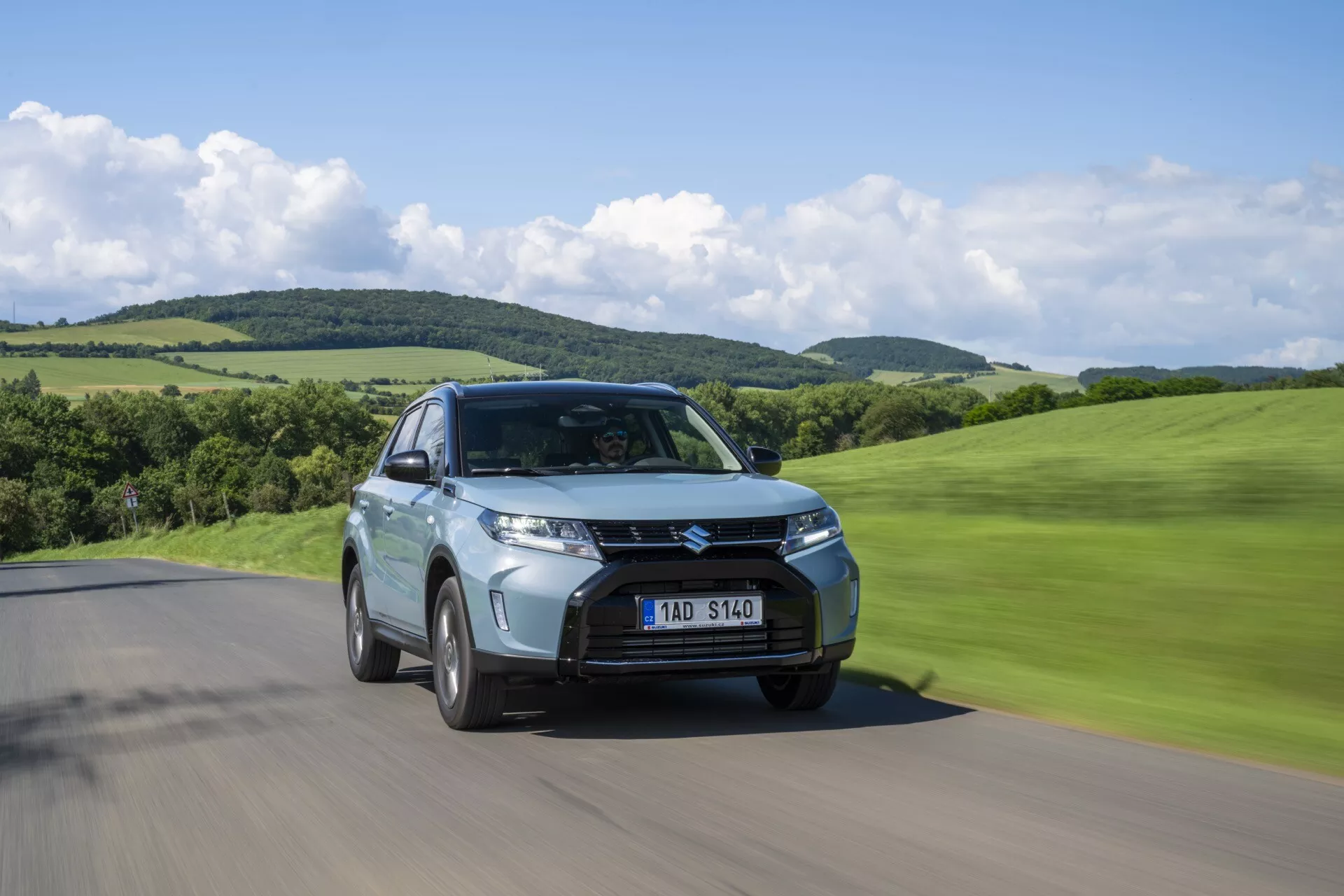 Záber z nízkeho uhla ukazujúci druhú generáciu Suzuki Vitara 2024 facelift na ceste, predný trojštvrtinový pohľad zobrazujúci mriežku, svetlomety a pohybovú rozmazanosť v zelenej, kopcovitej krajine.