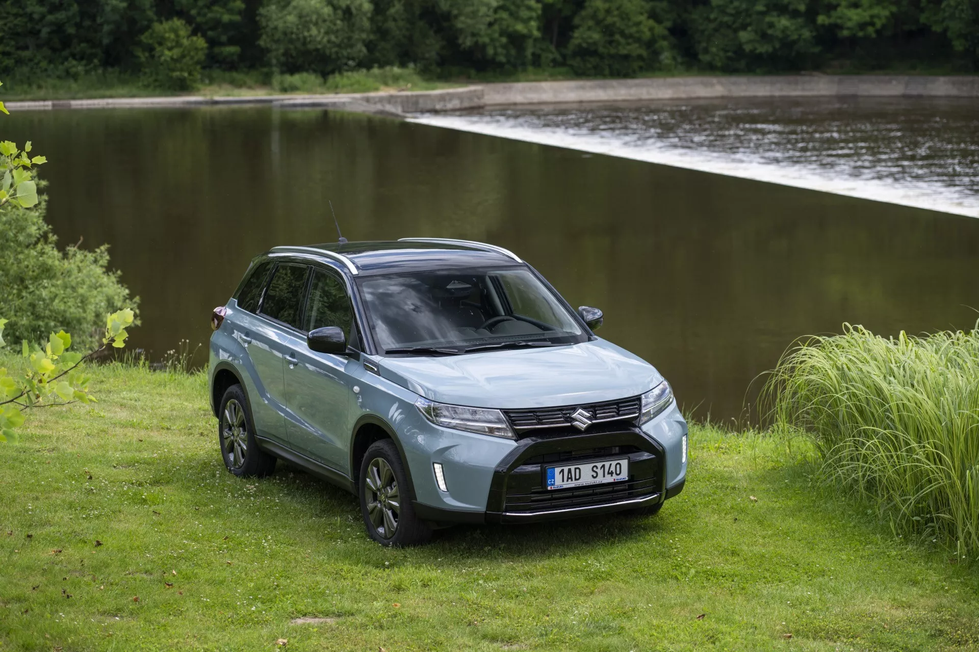 Na obrázku je znázornené SUV Suzuki Vitara 2024 (druhá generácia, druhý facelift) v pokojných vonkajších prostrediach blízko vodnej plochy. Zachytené z trojštvrťového predného pohľadu, SUV má viditeľné moderné LED svetlá, mriežku chladiča a predný nárazník. Sochársky vytvarovaná kapota a sklopené čelné sklo prispievajú k aerodynamickému profilu. Na jednej strane vidno dizajn so štyrmi dverami, uzavreté zadné úložné miesto, disky z ľahkej zliatiny a robustné terénne pneumatiky. Tlmená strieborná alebo šedá farba pridáva na elegancii. Nezpevnený prírodný terén okolo SUV podčiarkuje jeho schopnosti off-road a dobrodružný charakter.