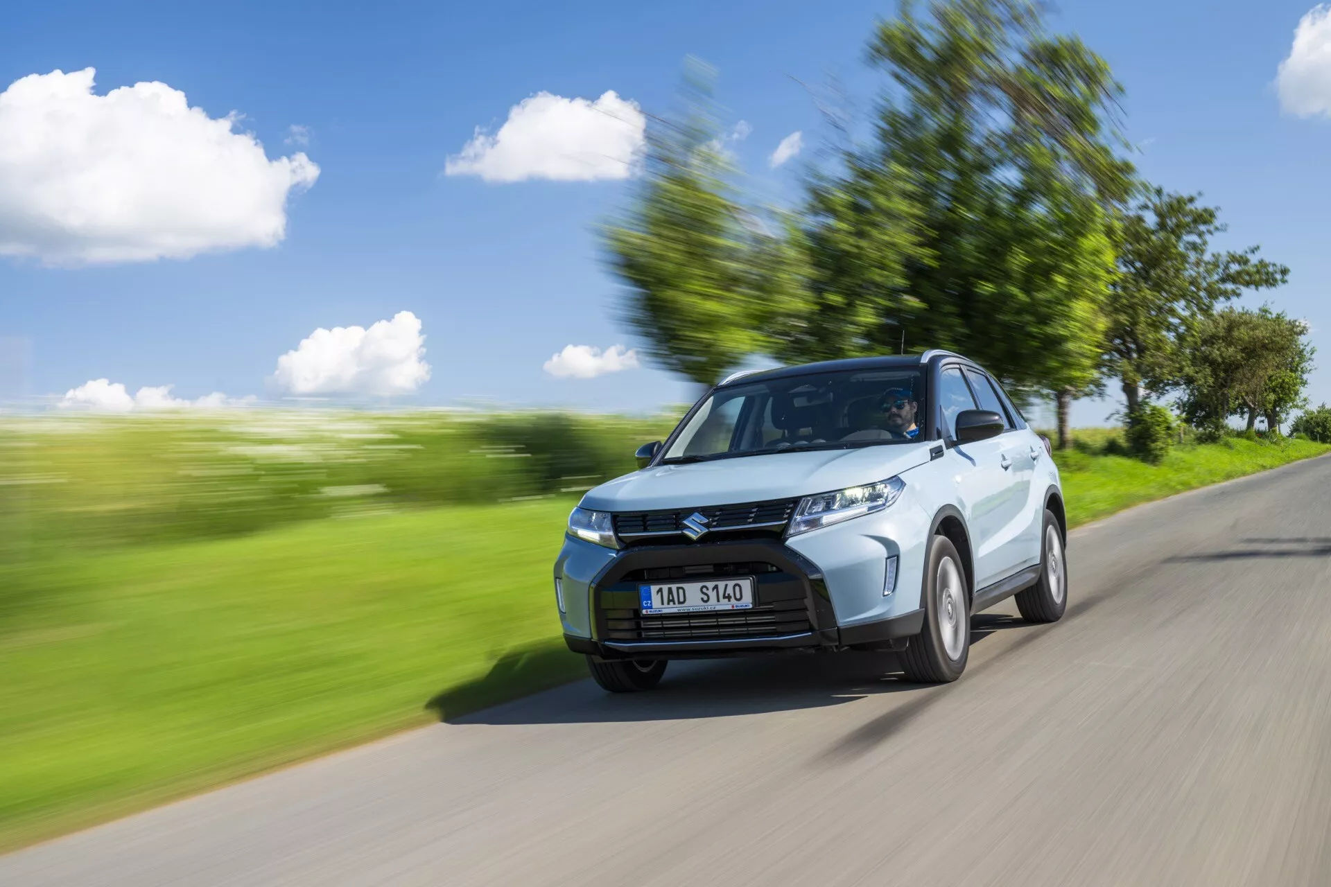 Dynamický záber Suzuki Vitara 2024 (2. generácia, facelift) zachytený z predného trojštvrťového pohľadu. Auto ide po ceste s rozmazanou zelenou krajinou v pozadí, čo naznačuje pohyb. Viditeľné prvky zahŕňajú kapotu, mriežku chladiča, svetlomety, predný nárazník a mierne natočené predné koleso. Strana vodiča, vrátane dverí a spätného zrkadla, je výrazne viditeľná. Pohybové rozmazanie a pruhy pozdĺž okolia zvyšujú pocit rýchlosti, zatiaľ čo auto zostáva zaostrené. Zreteľné sú kontúry, rezy na strane a dizajn kolies. Jasné denné svetlo s jasnou oblohou a tieňom naznačuje slnečné svetlo z horného ľavého smeru. Cez čelné sklo je vidieť postavu, pravdepodobne vodiča.