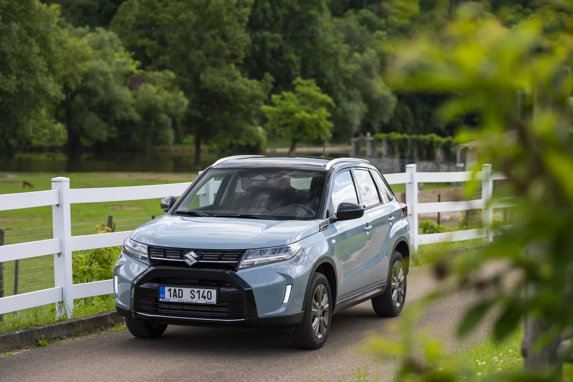 Svetlomodré Suzuki Vitara 2024 SUV v parku alebo vidieckom prostredí, zobrazené z predného trojštvrtinového uhla, čo ukazuje jeho sofistikovanú mriežku s emblémom, ostré LED svetlá, aerodynamické línie kapoty a viacramenné zliatinové kolesá. Predná ŠPZ vozidla je rozmazaná a spätné zrkadlá môžu mať integrované smerovky. Fotografia zachytáva auto s plytkou hĺbkou ostrosti, zdôrazňujúc jeho moderný dizajn, zatiaľ čo pozadie zostáva mierne rozmazané.