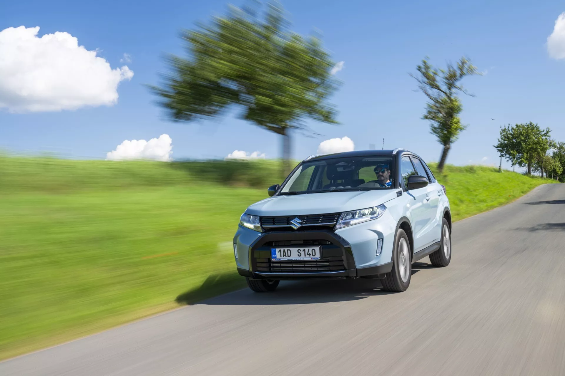 Dynamický záber na Suzuki Vitara 2024 (2. generácia facelift), ktorý ukazuje jej moderný dizajn a rýchlosť. Zachytené z pohľadu prednej ľavej štvrtiny, obraz zdôrazňuje ostré svetlomety, výraznú prednú mriežku a štandardné zliatinové kolesá. Efekt rozmazania pohybu na pozadí a kolesách zvýrazňuje rýchly pohyb auta, ako je vidieť na rozmazanej ceste s rastlinami a stromami.