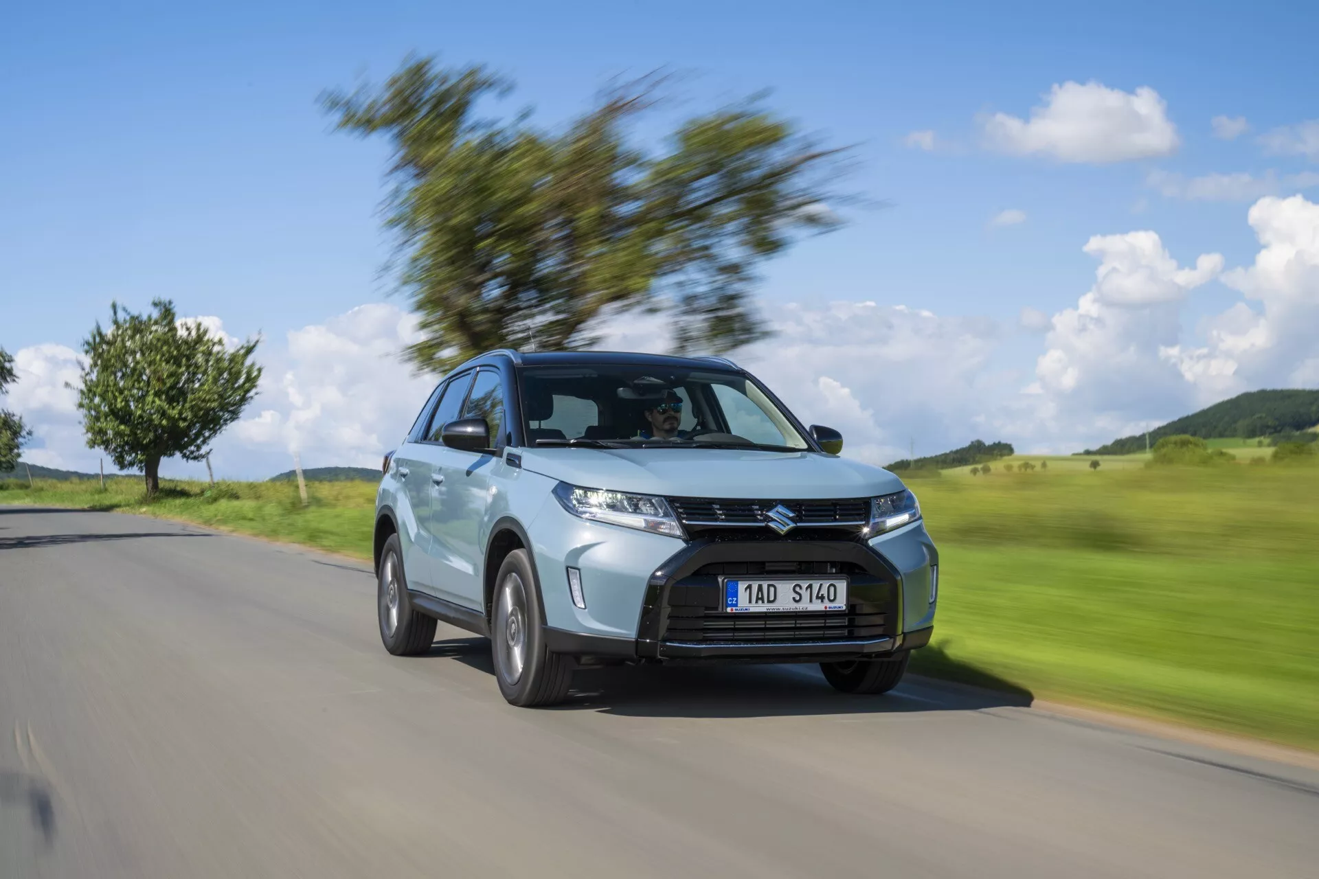 Dynamický obrázok Suzuki Vitara 2024 (2. generácia, facelift 2) v pohybe, zobrazujúci predný trojštvrtinový uhol. Auto je ostro zaostrené, zatiaľ čo pozadie je rozmazané pohybom, zvýrazňujúc jeho svetlomety, výraznú masku chladiča, zliatinové kolesá, kompaktný SUV karosériu, metalický náter a aerodynamický dizajn. Ideálne na automobilové výstavy a propagačné použitie.