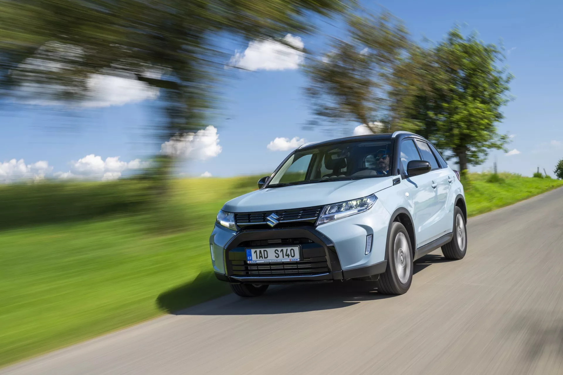 Svetlomodrý kompaktný SUV Suzuki Vitara 2024 (2. generácia facelift) v pohybe na vidieckej ceste. Obraz zachytáva prednú časť automobilu, ukazuje prednú mriežku, svetlomety, časť kapoty, čelné sklo a stranu vodiča vrátane predného blatníka, časť zadných dverí a časť zadného kolesového oblúka. Auto je ostré oproti rozmazanému pozadiu a ceste, čo naznačuje rýchlosť. Kolesá sú mierne rozmazané, čo naznačuje rotáciu. Osvetlené jasným prirodzeným svetlom počas slnečného dňa, vrhajúc tiene na spodnú časť vozidla, obrázok zdôrazňuje dizajnové prvky, ako sú záhyby na kapote, dizajn predného nárazníka a vzor mriežky. LED svetlomety sú viditeľné, čo môže naznačovať moderné funkcie ako parkovacie senzory. ŠPZ je viditeľná bez citlivých informácií.