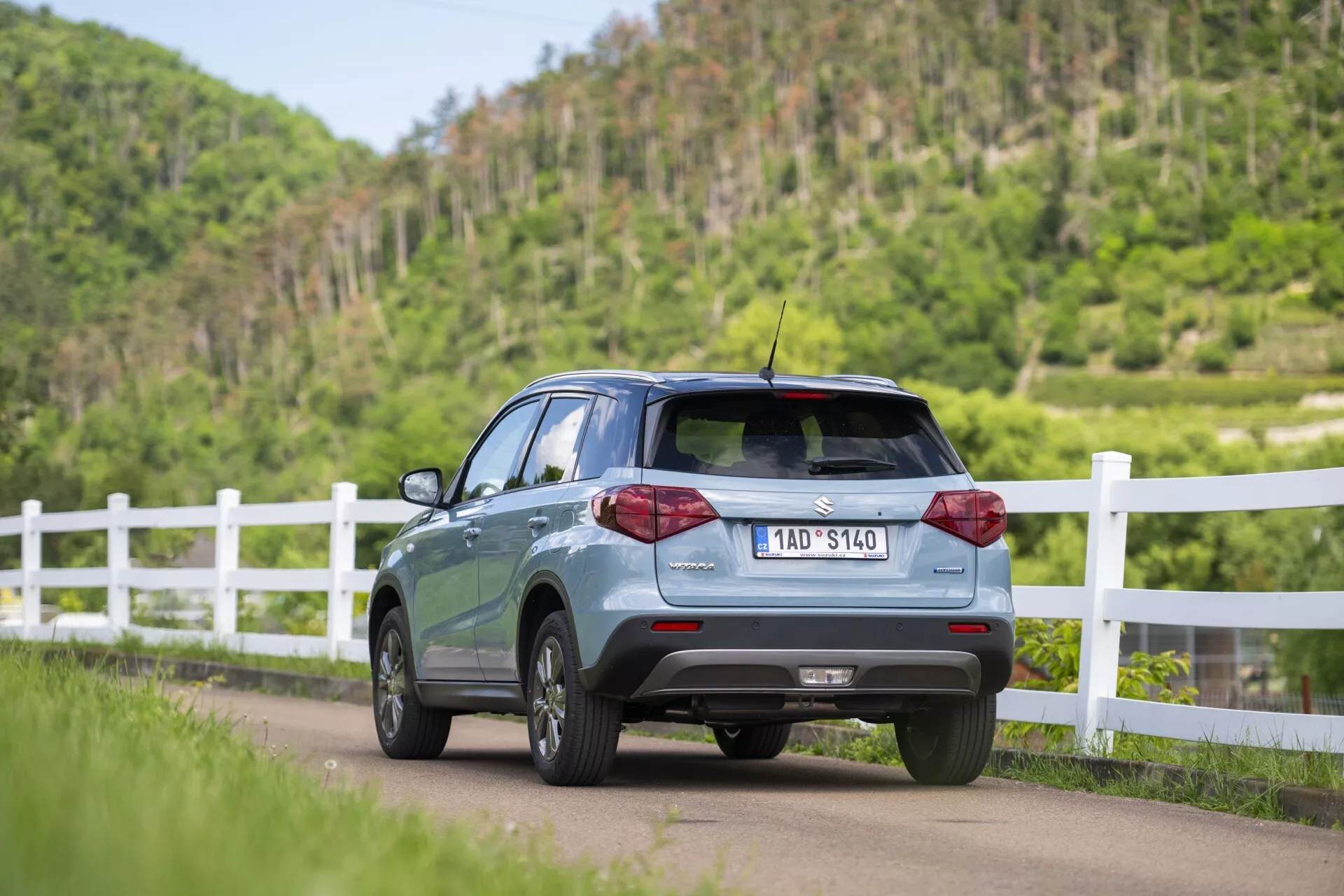 Obrázok zobrazuje Suzuki Vitara 2024 (2. generácia, facelift) z uhla troch štvrtín zozadu, zdôrazňujúci profil zadnej pravej strany umiestnený na ceste a mierne naklonený doľava. Viditeľné prvky zahŕňajú zadné dvere, zadné okno, čiastočné dvere a okná na pravej strane pre pasažierov, LED zadné svetlá, parkovacie senzory na zadnom nárazníku, strešný spoiler a krátku strešnú anténu. Auto má zliatinové kolesá a viditeľnú registračnú značku. Je zasadené do vidieckeho prostredia s bielym plotom a bujnou zeleňou.