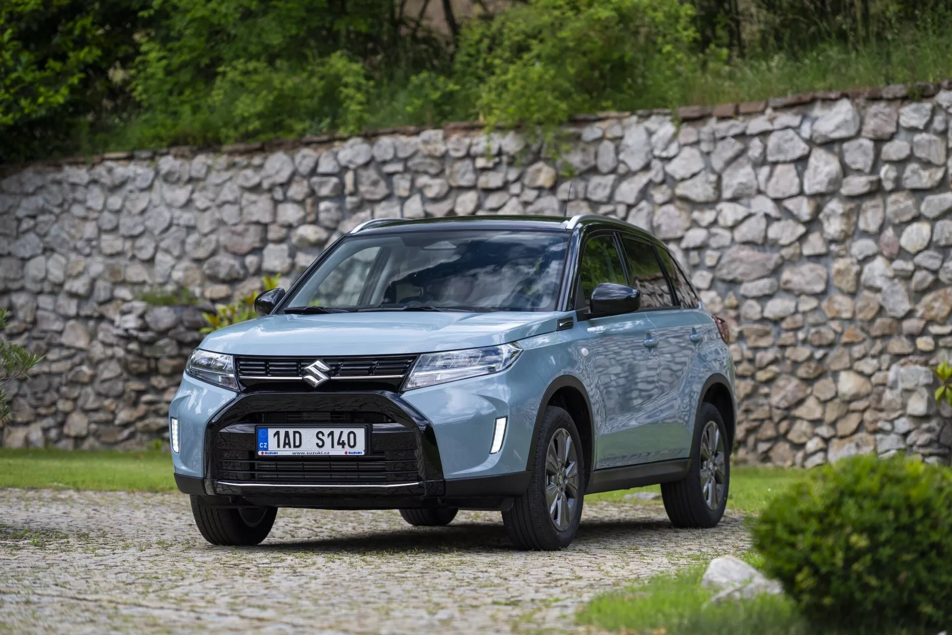 Vysokokvalitná fotografia automobilu Suzuki v exteriéri, zachytená z uhla tri štvrtiny prednej strany blízko zelene alebo parku. Pohľad zahŕňa prednú masku so zvýrazneným znakom Suzuki, moderné svetlomety pravdepodobne s LED technológiou, hmlové svetlá v nárazníku a profil vodičovej strany s viditeľnými kolesami, dverami a zrkadlami. Auto stojí na štrkovej ploche s kamenným múrom v pozadí, zdôrazňujúc prírodnú, rustikálnu atmosféru. Jemne osvetlená, pravdepodobne zamračená scéna má plytkú hĺbku ostrosti, rozmazávajúc pozadie, aby sa zamerala na dizajn a technologické detaily auta.