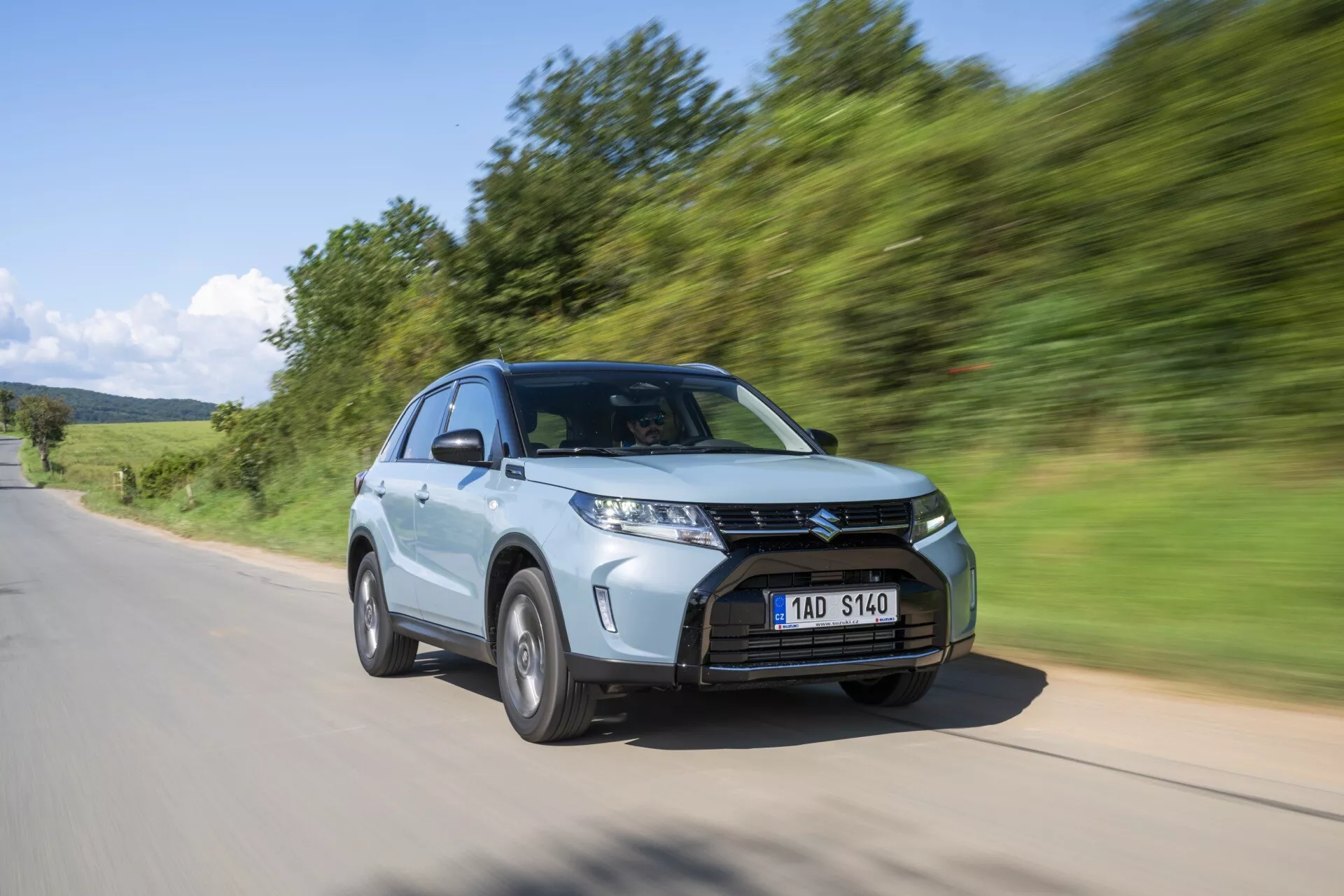 Moderné SUV, Suzuki Vitara 2024 (druhá generácia, facelift 2) zachytené v pohybe z nízkeho a predného trojštvrťového uhla. Obrázok zvýrazňuje výraznú prednú časť vozidla vrátane výrazne navrhnutého grilu s horizontálnymi lamelami, ostrých a uhlových LED svetiel a viacramenného hliníkového kolesa. SUV je zachytené pri jazde po asfaltovej ceste s prirodzeným zeleným pozadím, s rozmazaním pohybu, ktoré vyjadruje rýchlosť a dynamiku. Ďalšie vlastnosti zahŕňajú naklonenú kapotu, aerodynamické zrkadlá pre lepšiu aerodynamiku, vyššiu svetlú výšku a priestranný interiér, všetko zvýraznené metalickým lakom.