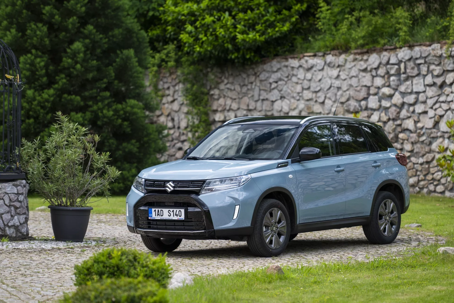 Šikmý predný bočný pohľad na moderné SUV Suzuki Vitara 2024 zaparkované vonku v rezidenčnej alebo parkovej oblasti s dobre udržiavaným trávnikom a kamenným múrom v pozadí. Obrázok, vytvorený zo strednej vzdialenosti, ukazuje prednú mriežku s horizontálnymi pruhmi s logom spoločnosti, elegantné LED svetlá s dennými svietidlami, sochársky tvarovaný nárazník s integrovanými hmlovými svetlami, hliníkové kolesá, integrované smerové svetlá v bočných zrkadlách, kovový povrch laku, dynamické línie karosérie, tónované okná, strešné nosiče a strešnú anténu, zdôrazňujúc jeho moderný dizajn a vlastnosti.