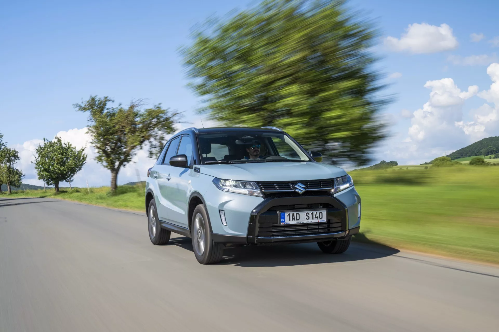 Dynamický obraz strieborného hatchbacku Suzuki Vitara 2024 v pohybe zachytený z trojštvrťového predného uhla, ktorý zdôrazňuje moderný dizajn s výraznou mriežkou, hranatými svetlometmi, klesajúcou strechou a kolesami v pohybe s rozmazaným pozadím vyjadrujúcim rýchlosť.