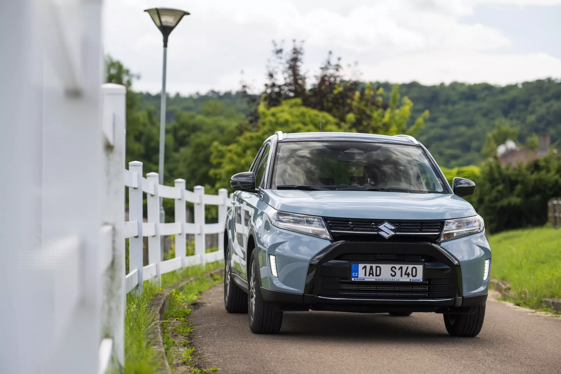 Denná scéna vonka zobrazujúca Suzuki Vitara 2024 (2. generácia facelift) zachytený z predného trojštvrťového pohľadu. Obrázok zdôrazňuje dizajnové prvky vozidla vrátane výraznej prednej masky s horizontálnym mriežkovým dizajnom a centrálnym znakom, moderné LED alebo HID svetlomety, zapustené hmlovky v spodnom nárazníku a svetlo modrej alebo sivej farby. Auto ide po úzkej ceste blízko bieleho plotu s mierne rozmazaným pozadím, čo naznačuje malú hĺbku ostrosti. Viditeľné prvky zahŕňajú taktiež zliatinové kolesá, bočné zrkadlá so zabudovanými smerovkami a čiastočne viditeľný predný nárazník s centrálnym prívodom vzduchu.