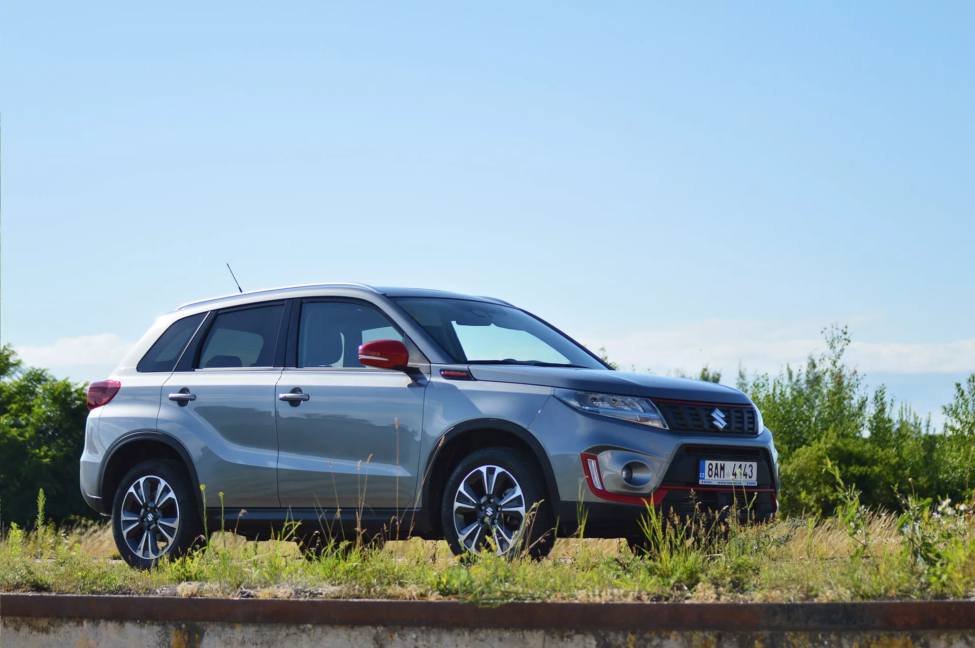 Bočný profil strieborného kompaktného SUV Suzuki Vitara 2018, zaparkovaného paralelne vedľa železničnej trate za slnečného dňa. Sú jasne viditeľné dizajnové línie, kolesá z ľahkých zliatin s viaclúčovým dizajnom, metalízový lak, zatvorené dvere a okná, tvar svetlometov, bočné zrkadlo s potenciálne integrovanými smerovkami a odznak úrovne výbavy, čím sa zdôrazňuje dizajn a technologické prvky vozidla.