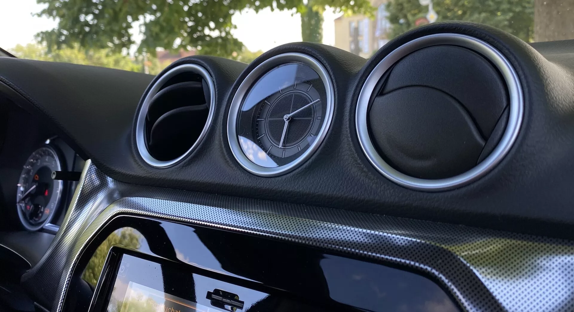 Detailný pohľad na interiér Suzuki Vitara 2018 so zameraním na centrálny palubný panel. Palubný panel obsahuje tri kruhové kovovo vyzerajúce vetracie otvory s emblémom výrobcu, dotykový infotainment systém s lesklým čiernym rámom, materiál vzhľadu karbónových vlákien a ambientné osvetlenie pre zlepšenie estetiky.