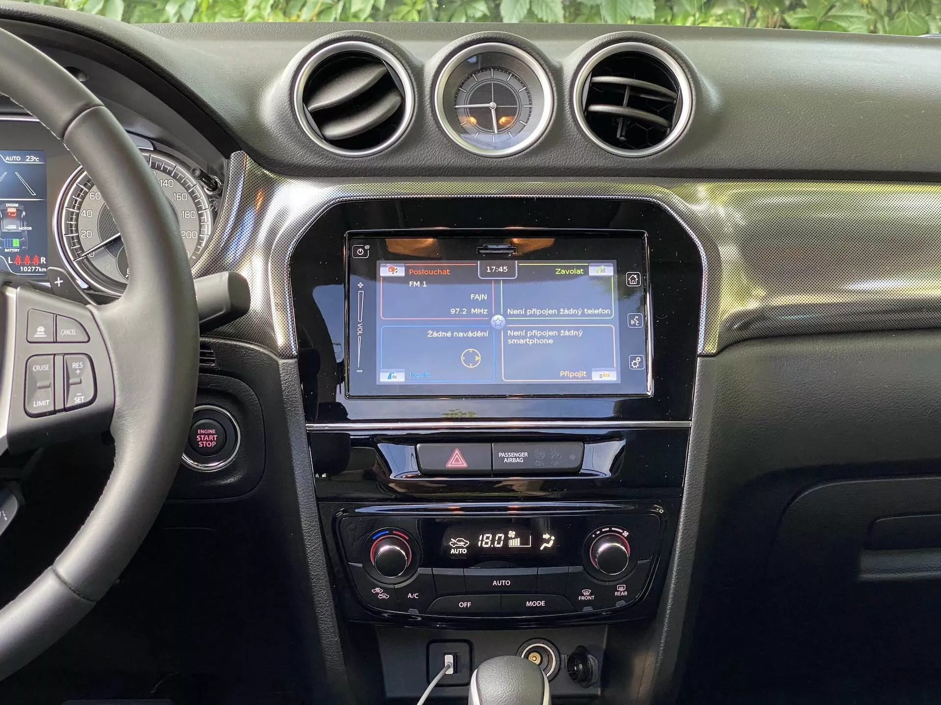 Interiérový pohľad na Suzuki Vitara 2018 so zameraním na stredovú prístrojovú dosku. Sekcia obsahuje čiastočne viditeľný volant vľavo s niekoľkými ovládacími tlačidlami, pravdepodobne pre ovládanie audia, handsfree volania a tempomatu. Stredová konzola zobrazuje dotykovú obrazovku infotainmentu, pravdepodobne zobrazujúcu navigáciu alebo menu, obklopenú vertikálnymi ventilátormi a zakončenú tromi kruhovými vetracími otvormi. Pod dotykovou obrazovkou je rad tlačidiel a ovládacích prvkov na rýchly prístup k nastaveniam infotainmentu a vozidla. Ovládacie prvky klímy sú umiestnené nižšie, čo naznačuje, že auto má dvojzónový klimatizačný systém. Prístrojová doska má mäkký materiál s viditeľným prešívaním a medzi centrálnymi kruhovými ventilátormi je umiestnené guľaté analógové hodiny. Viditeľnosť sa sústreďuje na prvky prístrojovej dosky, bez viditeľných okien alebo čelného skla.