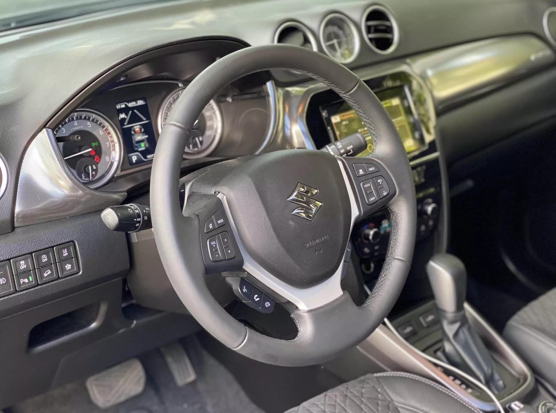Interiér Suzuki Vitara 2018, zachytávajúci perspektívu vodiča. Detail koženého viacfunkčného volantu s emblémom, ovládacími prvkami pre zvuk, hlasové ovládanie a tempomat s kovovými akcentmi. Za ním sa nachádza prístrojová doska s analógovými číselníkmi pre rýchlomer a otáčkomer a digitálnou obrazovkou pre informácie o vozidle, naznačuje, že auto je vypnuté alebo v režime príslušenstva. Stredová konzola ukazuje kovovú alebo chrómovú hromadu s dotykovým infotainment systémom, tlačidlami a gombíkmi pre ovládanie klímy a vozidla. Automatická radiaca páka s koženými akcentmi naznačuje rôzne režimy jazdy. Interiér kombinuje kožu a kartáčovaný kov, čo odráža moderný dizajn. Predpokladané pokročilé funkcie zahŕňajú Bluetooth, navigáciu, asistentov parkovania a systémy asistencie vodiča.