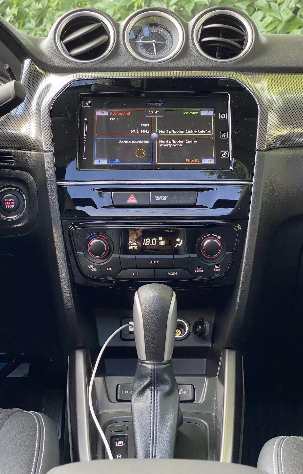 Podrobný pohľad na interiér Suzuki Vitara 2018, zdôrazňujúci stredovú konzolu a prístrojovú dosku z pohľadu predných sedadiel. Obrázok zvýrazňuje dotykovú obrazovku infotainmentu zobrazujúcu menu, dvojzónový klimatizačný systém s digitálnymi ukazovateľmi a páku automatickej prevodovky. Interiér ukazuje tmavé farby s mixom matných a lesklých textúr, ilustrujúci kvalitu materiálu a modernú automobilovú technológiu.