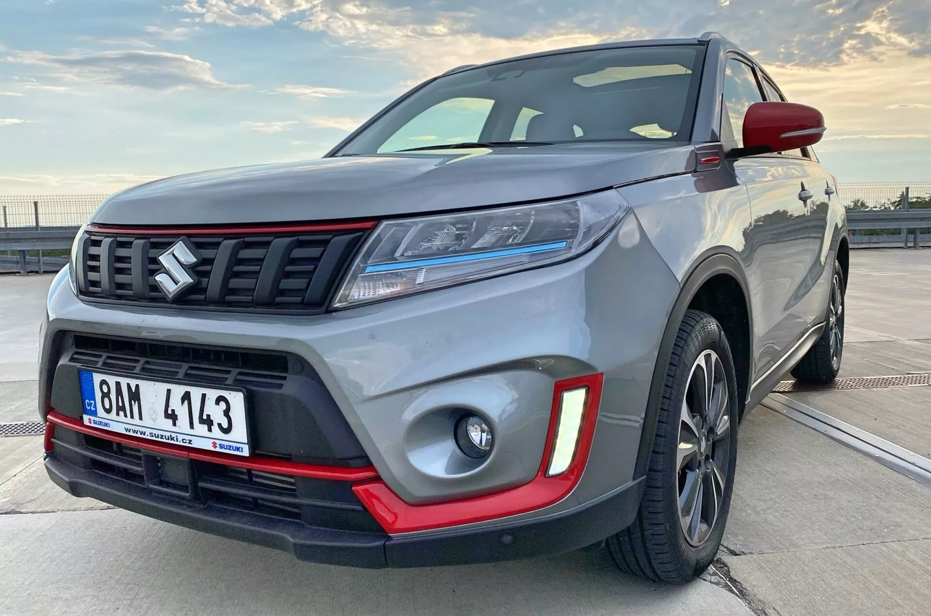 Blízky pohľad na moderné kompaktné SUV z bočnej prednej strany, šikmo ukazujúc časť bočného profilu. Obrázok zdôrazňuje robustnosť vozidla s viditeľnými detailmi ako predný nárazník, mriežka chladiča s horizontálnymi lištami a emblémom, elegantné LED svetlomety, hmlovky s červenými vložkami, zliatinové kolesá a svalnaté blatníky. Tmavé zliatinové kolesá a kontrastné farebné prvky naznačujú prémiovú alebo športovú výbavu.