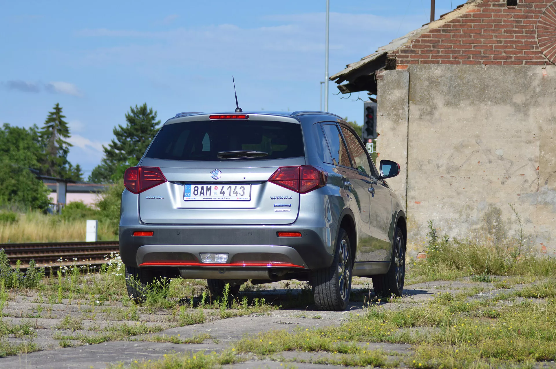 Metalická šedá Škoda Yeti, kompaktné SUV, zaparkované na zanedbanom pozemku s trávnatými a betónovými škvrnami, odfotené zo zadného trojštvrťového pohľadu. Obrázok ukazuje charakteristický zadný dizajn vozidla s konvenčnými zadnými svetlami, zadným stieračom, vysokou brzdovou svetlovou a časti viditeľným zadným spojlerom. Logo Škoda je umiestnené v strede na kufri, názov modelu 'Yeti' naľavo a znak '4x4' napravo, čo naznačuje pohon všetkých kolies. Auto má päťramenné zliatinové disky, vyzerá byť v dobrom stave a nachádza sa v priemyselnej oblasti s vyradenými železničnými koľajami a chátrajúcou štruktúrou v pozadí, pod prirodzeným slnečným svetlom.