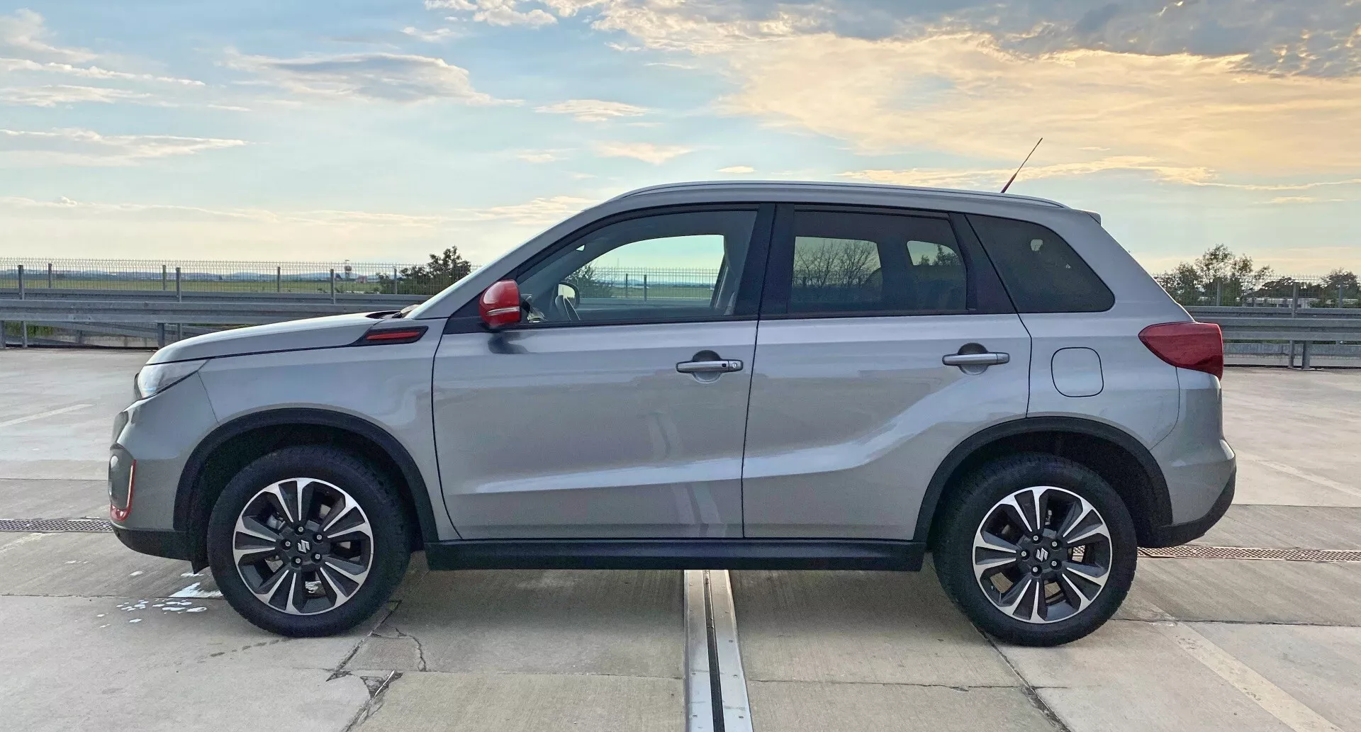 Bočný profil moderného SUV Suzuki Vitara 2018, odfotený počas denného svetla na priestrannom parkovisku. V popredí je stredná časť vozidla, ktorá ukazuje dĺžku a bočné dizajnové prvky, ako sú tvary dverí, bočné prahy a oblúky kolies. Prirodzené osvetlenie zvýrazňuje päť dverí, viacramenné zliatinové kolesá, strešné lišty, kontrastné kľučky a ozdobné prvky, anténu a možné tónované okná. SUV vykazuje vynikajúcu kvalitu s lesklým povrchom exteriéru.