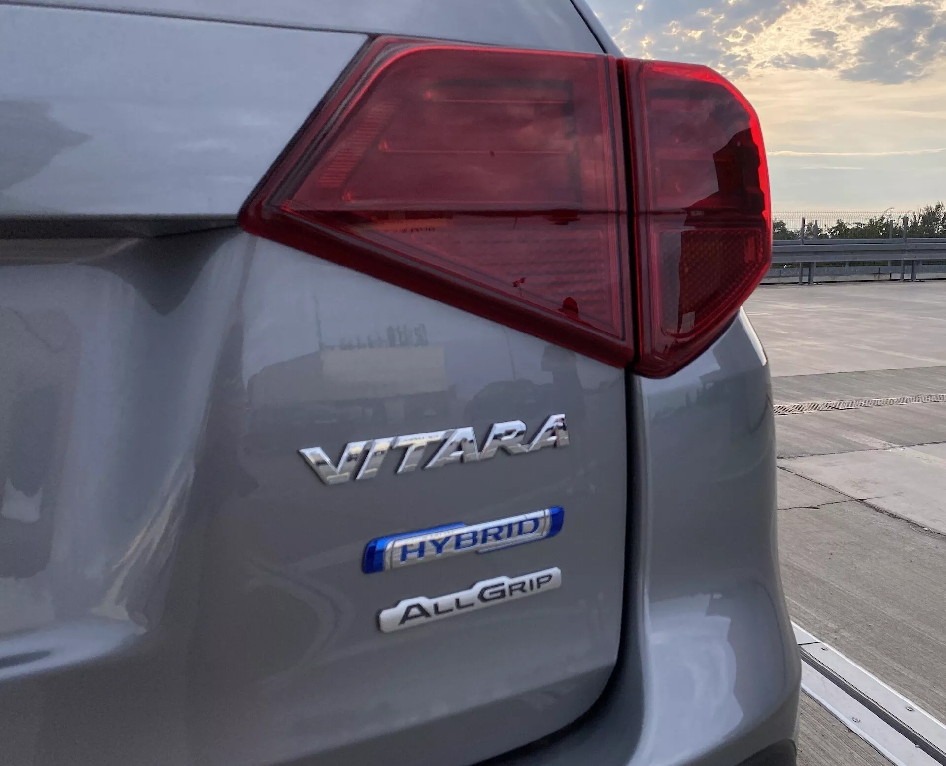 Detailný záber na ľavý zadný roh Suzuki Vitara 2018 ukazujúci zložitý dizajn zadného LED svetla a tri označenia uvádzajúce názov modelu 'VITARA', hybridnú technológiu 'HYBRID' a systém pohonu všetkých kolies 'ALLGRIP', zachytené pri úsvite alebo súmraku na otvorenom parkovisku.