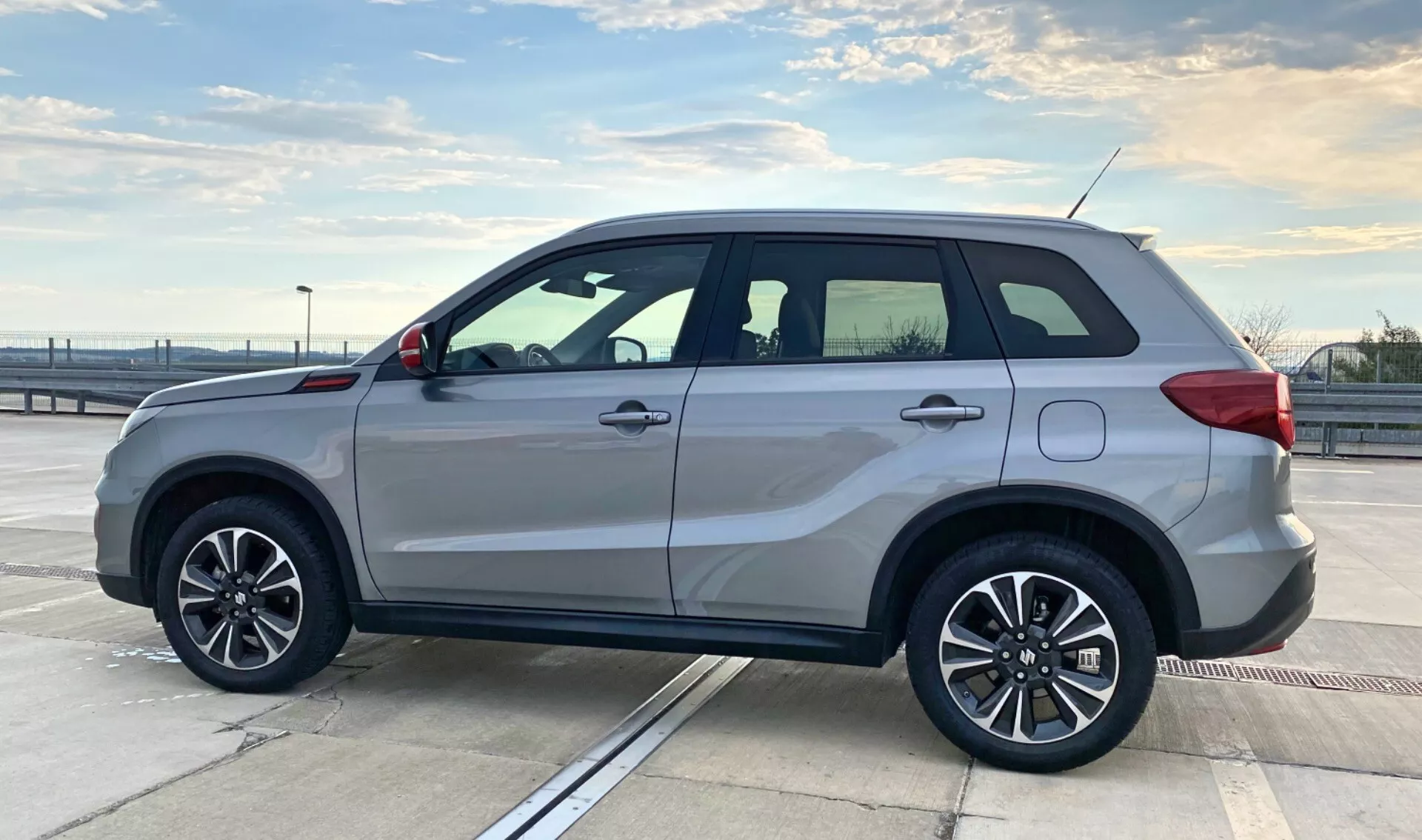 Bočný profilový pohľad na Suzuki Vitara 2018 zaparkovanú na hornom poschodí parkovacej konštrukcie pod jasnou oblohou. SUV má pätirapenné zliatinové kolesá, strieborný lak, integrované smerovky na bočných zrkadlách, strešné nosiče, dverové rukoväte vo farbe karosérie, jemný postranný nášľap a tónované okná s chrómovým lemovaním. Dizajn vozidla zdôrazňuje športovú eleganciu a úžitkovosť s hranatou karosériou pre maximalizáciu vnútorného priestoru. Vonkajšie prostredie a kompozícia zvýrazňujú štýl a stavbu vozidla, vhodné pre kategórie súvisiace s vozidlami.