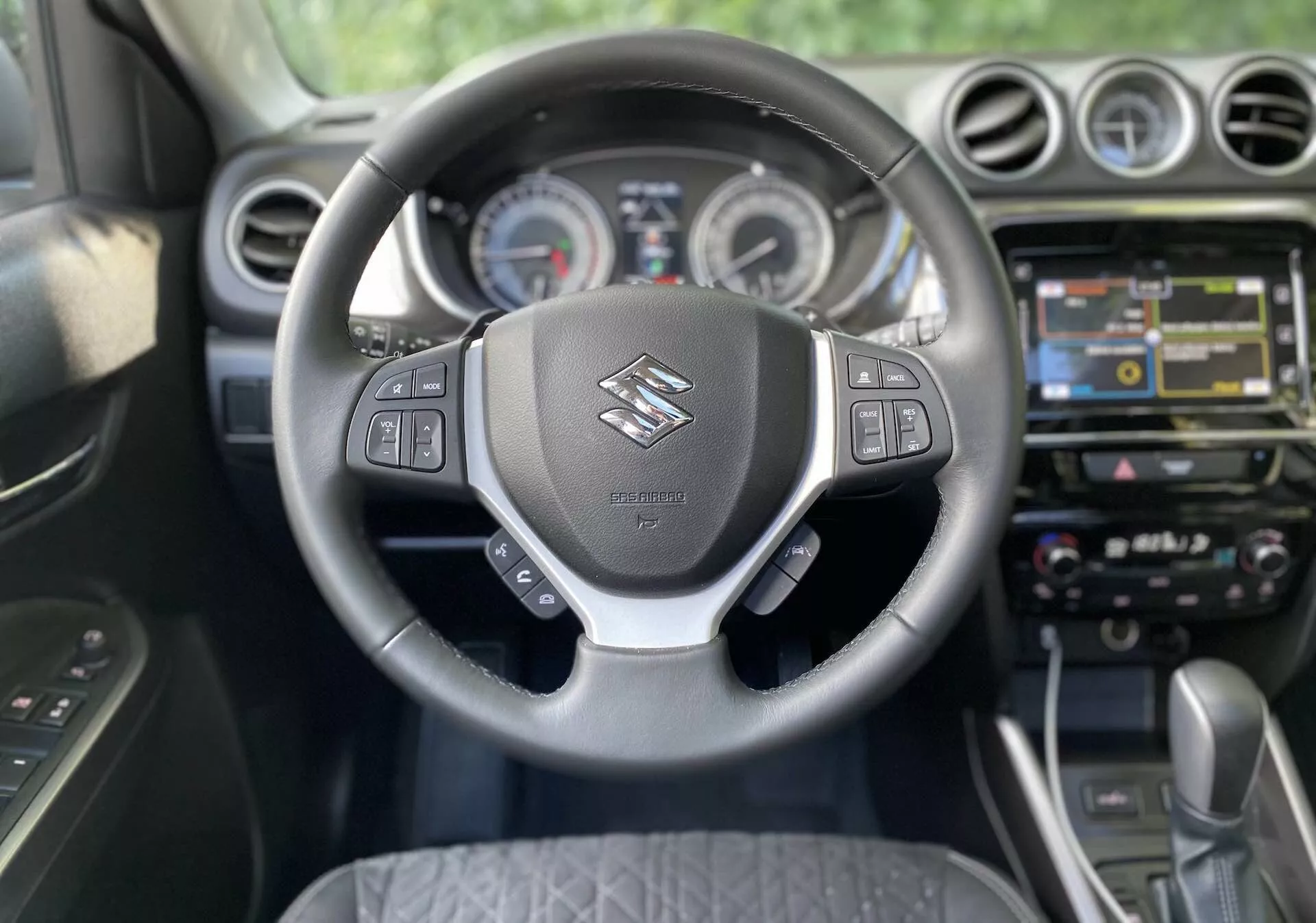 Pohľad do interiéru Suzuki Vitara 2018 z perspektívy vodiča. Hlavné zameranie je na volant s logom Suzuki a multifunkčnými ovládačmi pre audio, hlasové príkazy, tempomat a správu displeja informácií o vozidle. Za volantom sú viditeľné analógové ciferníky a centrálny displej prístrojovej dosky. Stredová konzola obsahuje dotykový infotainment systém s navigáciou, multimédiami a možnosťami pripojenia. Nižšie sú prítomné tlačidlá a otočné ovládače pre dvojzónovú klimatizáciu. Auto je vybavené automatickou prevodovkou, čo naznačuje moderný a prípadne prémiový dizajn kabíny.