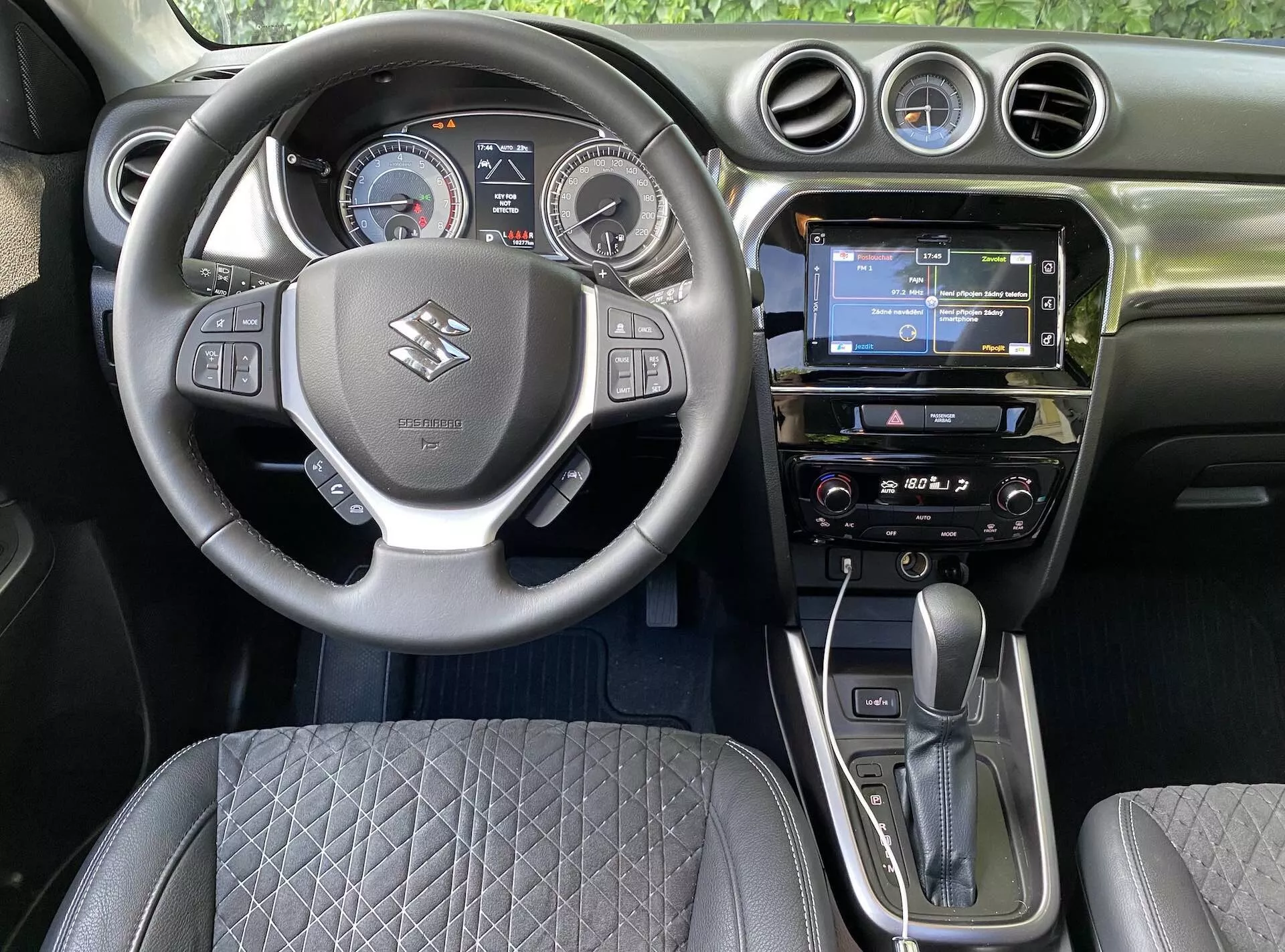 Pohľad na interiér Suzuki Vitara 2018 z miesta vodiča, ukazujúci stredový volant s logom značky, ovládacími tlačidlami pre hlasitosť, hlasové príkazy a pripojenie telefónu. Za volantom je digitálno-analógová prístrojová doska s rýchlomerom, otáčkomerom a centrálnym digitálnym displejom. Palubná doska obsahuje dotykovú infotainment obrazovku s GPS navigáciou, rádio a klimatizačný systém. Automatická radiaca páka a elektronická ručná brzda sú výrazne zobrazené, spolu s prešívaným dizajnom sedadla a kruhovými ventilačnými otvormi, zachytávajúce modernú automobilovú technológiu a ergonómiu dizajnu.